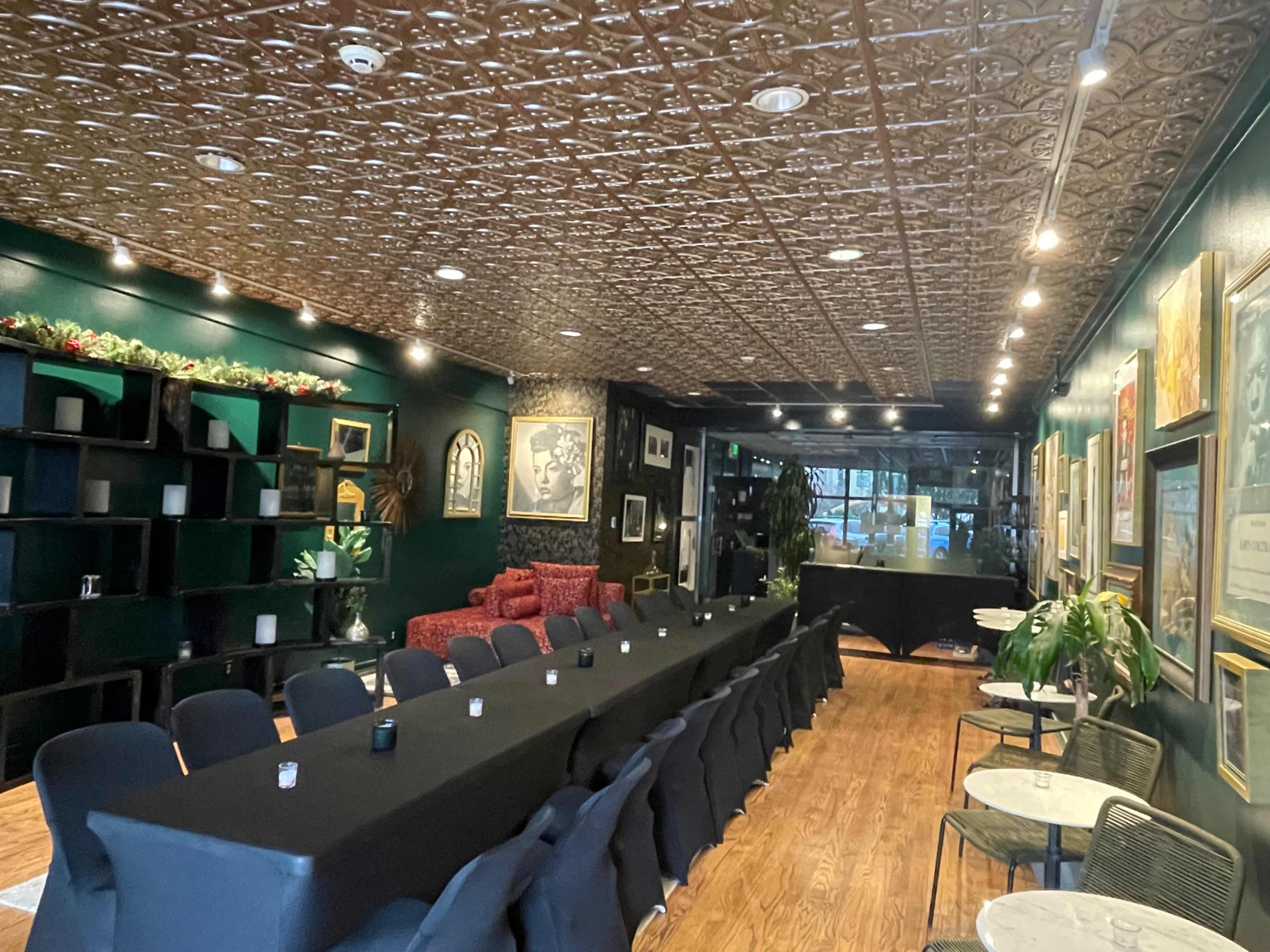 The image shows a spacious dining area with a long table set for a meal, surrounded by dark green walls, framed artwork, and elegant lighting.
