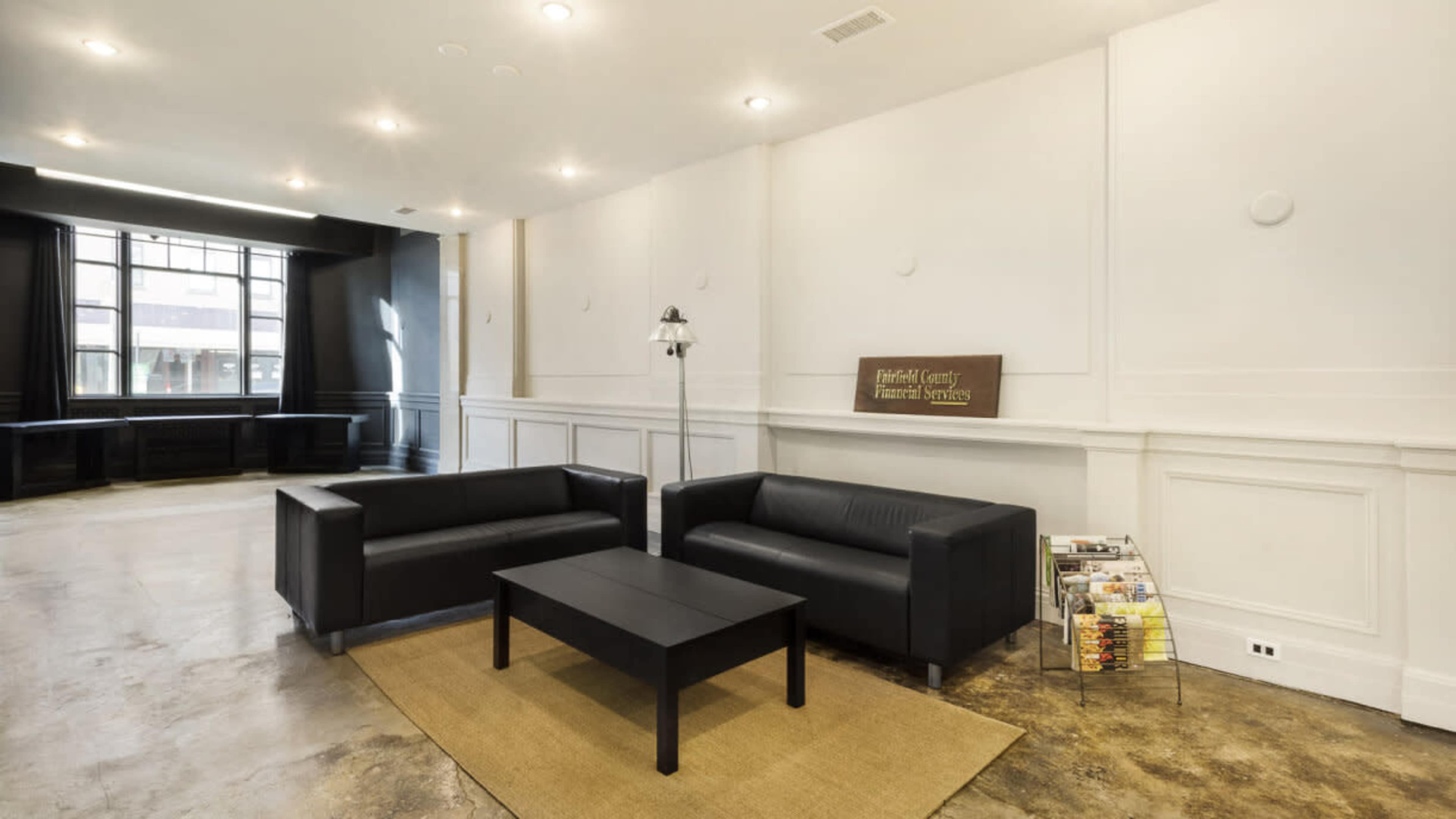 The image shows a modern waiting area with two black leather couches, a black coffee table, and a magazine rack, set against a light-colored wall and large windows.