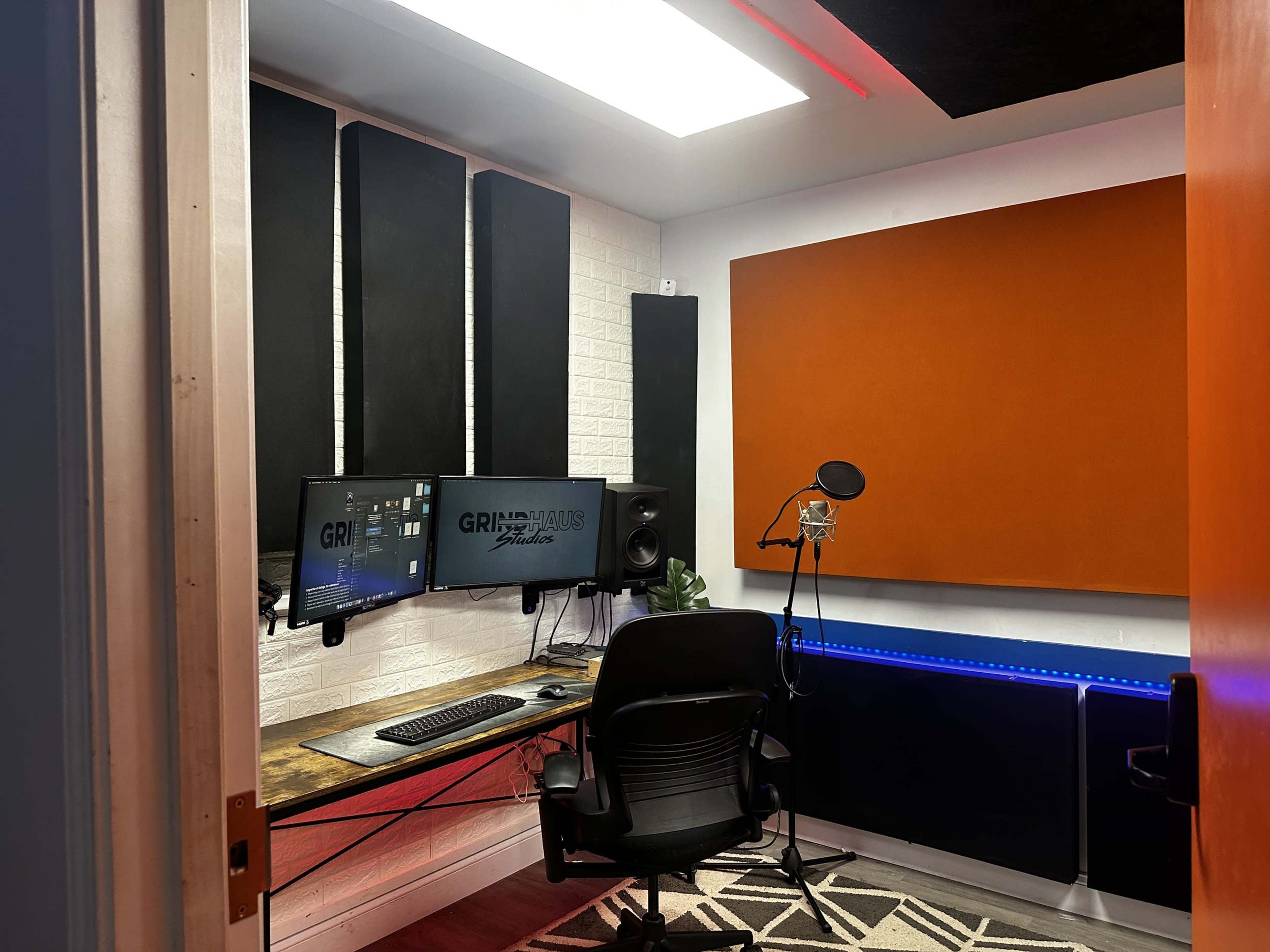 A modern music recording studio featuring two computer monitors, a microphone on a stand, and soundproofing panels on the walls.