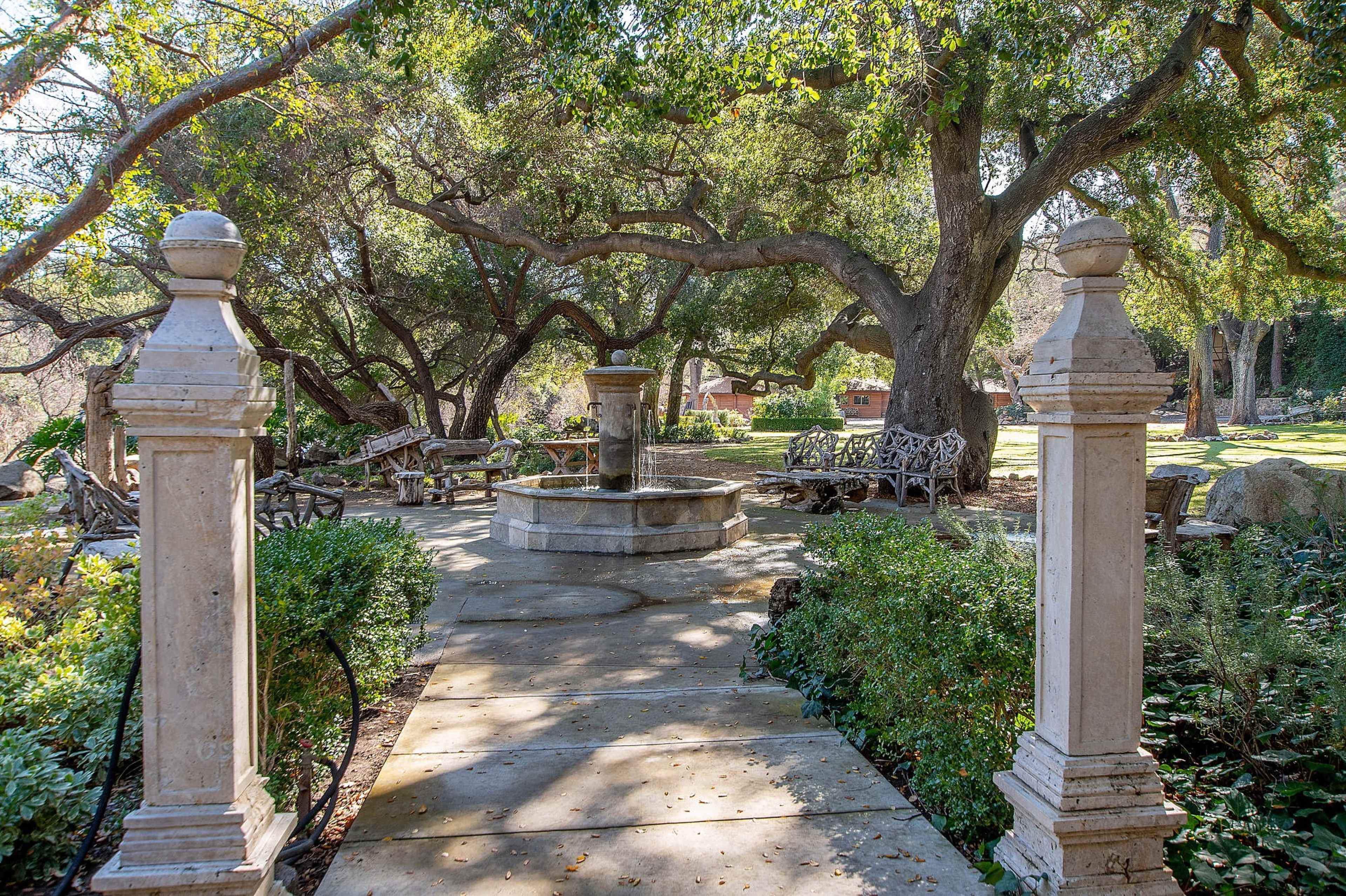 A stone pathway leads to a central fountain surrounded by lush greenery and shaded seating areas beneath large trees.
