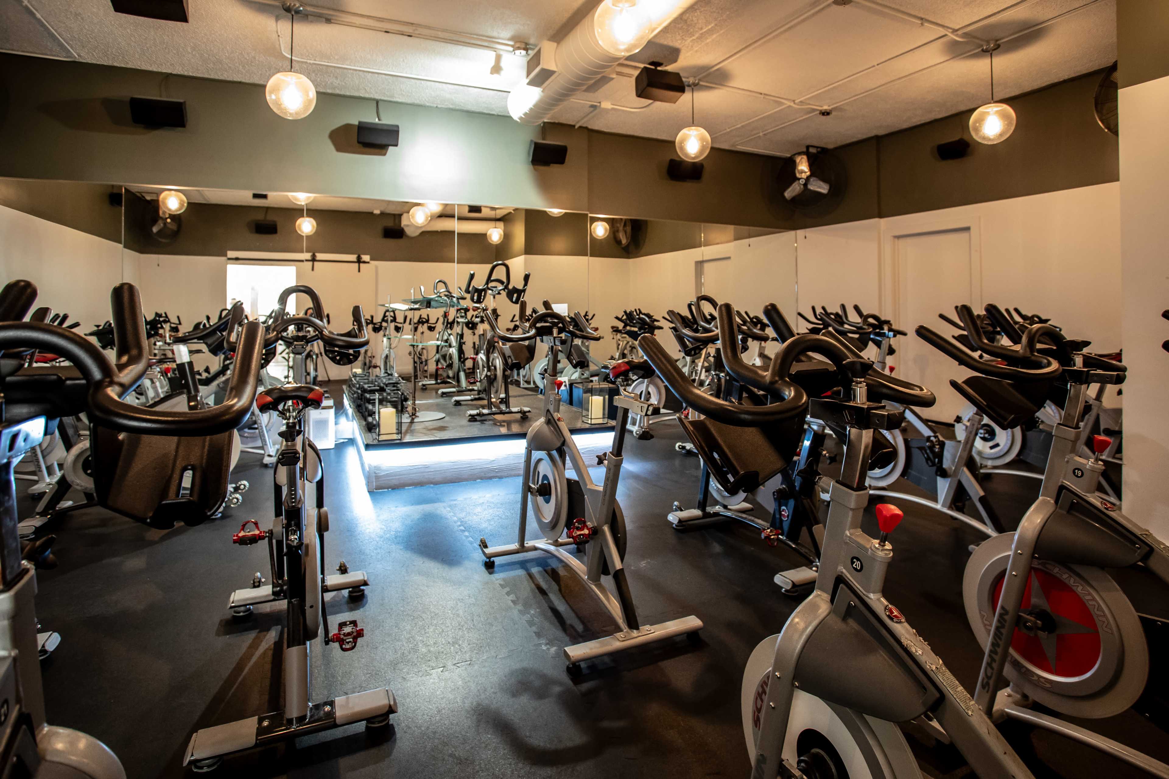 The image shows a spacious fitness studio filled with stationary bicycles arranged in rows, illuminated by overhead lights and natural light from windows.