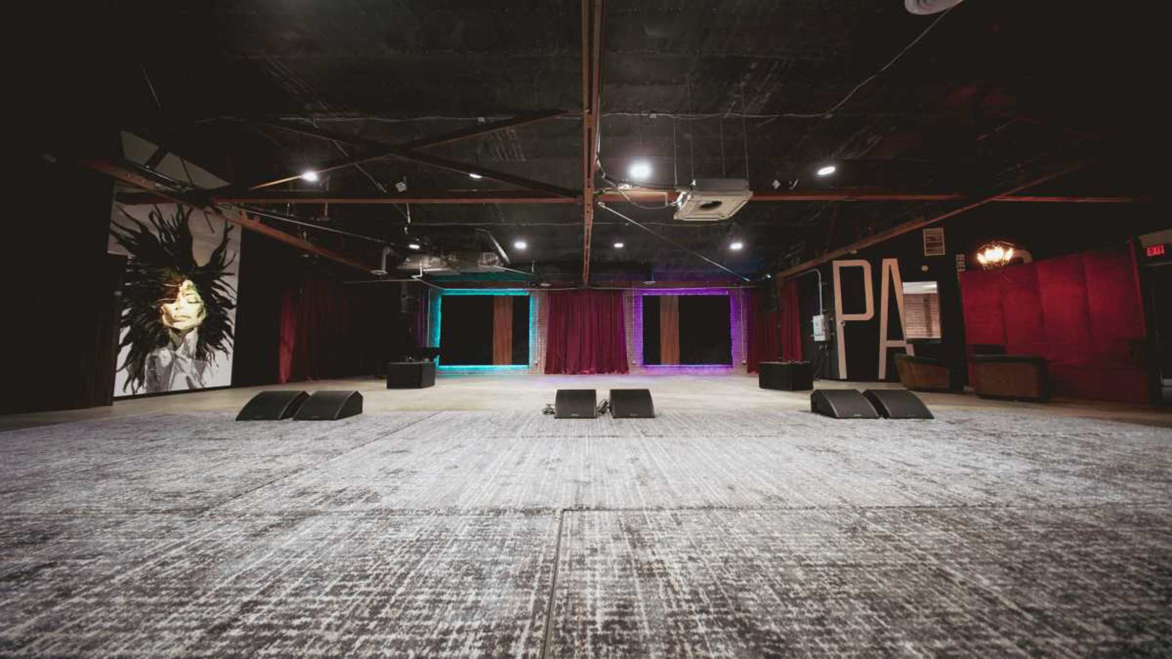 The image depicts an empty event space with a large, open floor, red curtains at the back, and colorful lighting options along the walls.