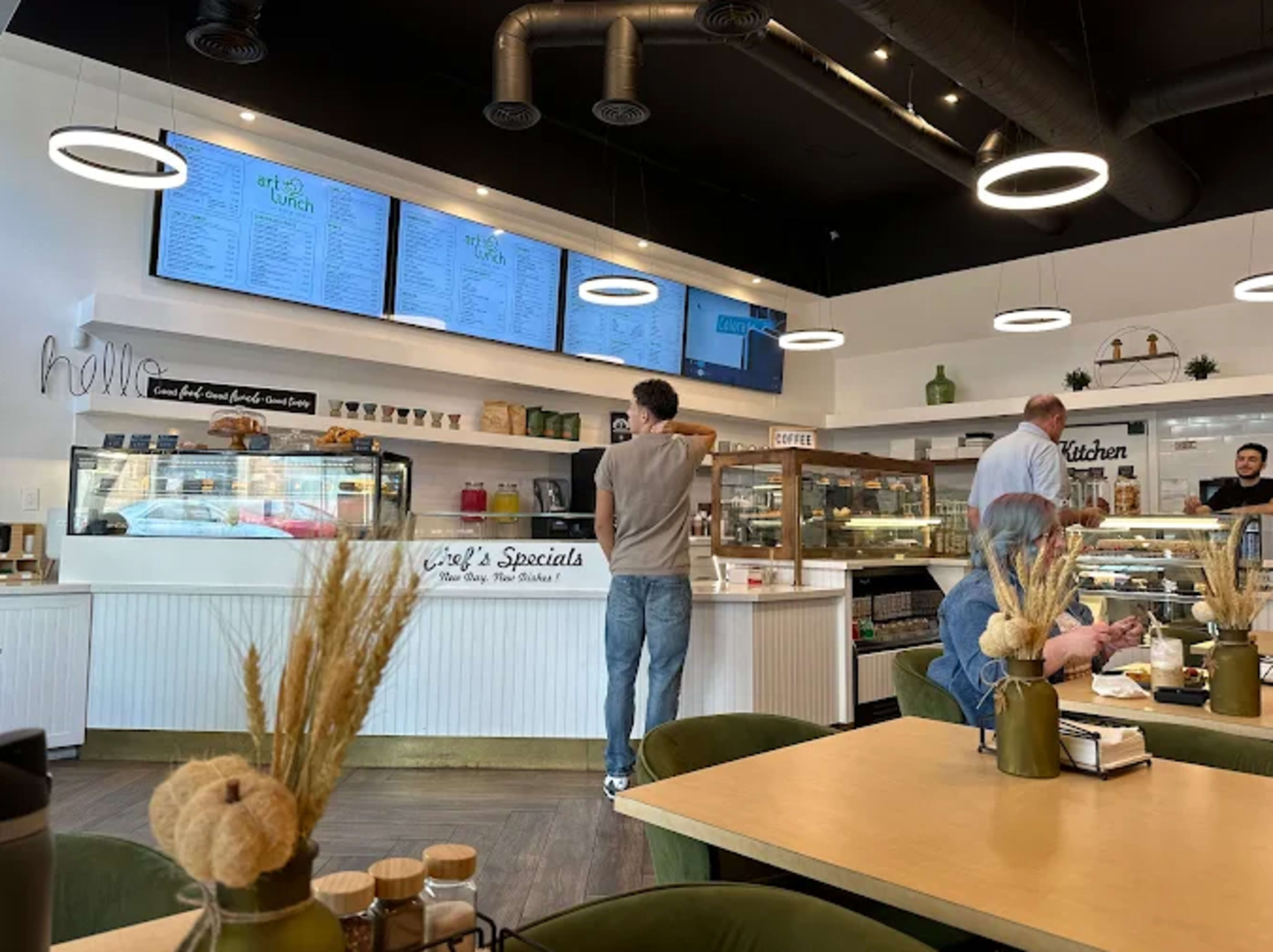A modern café interior features a display case with pastries, a menu on the wall, and customers seated at tables while one person is ordering at the counter.