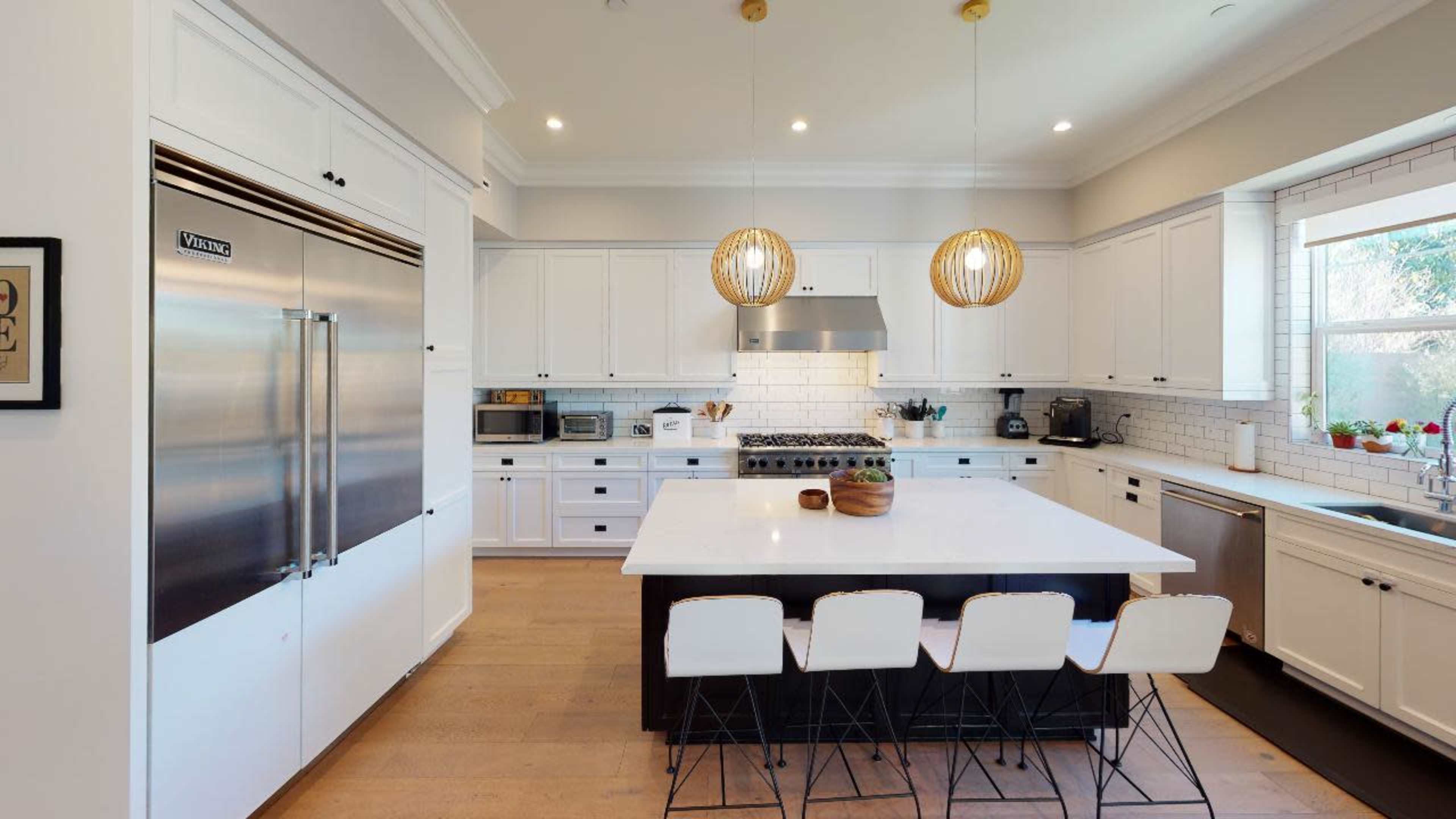 The image shows a modern kitchen featuring white cabinetry, a large island with four bar stools, stainless steel appliances, and pendant lighting.