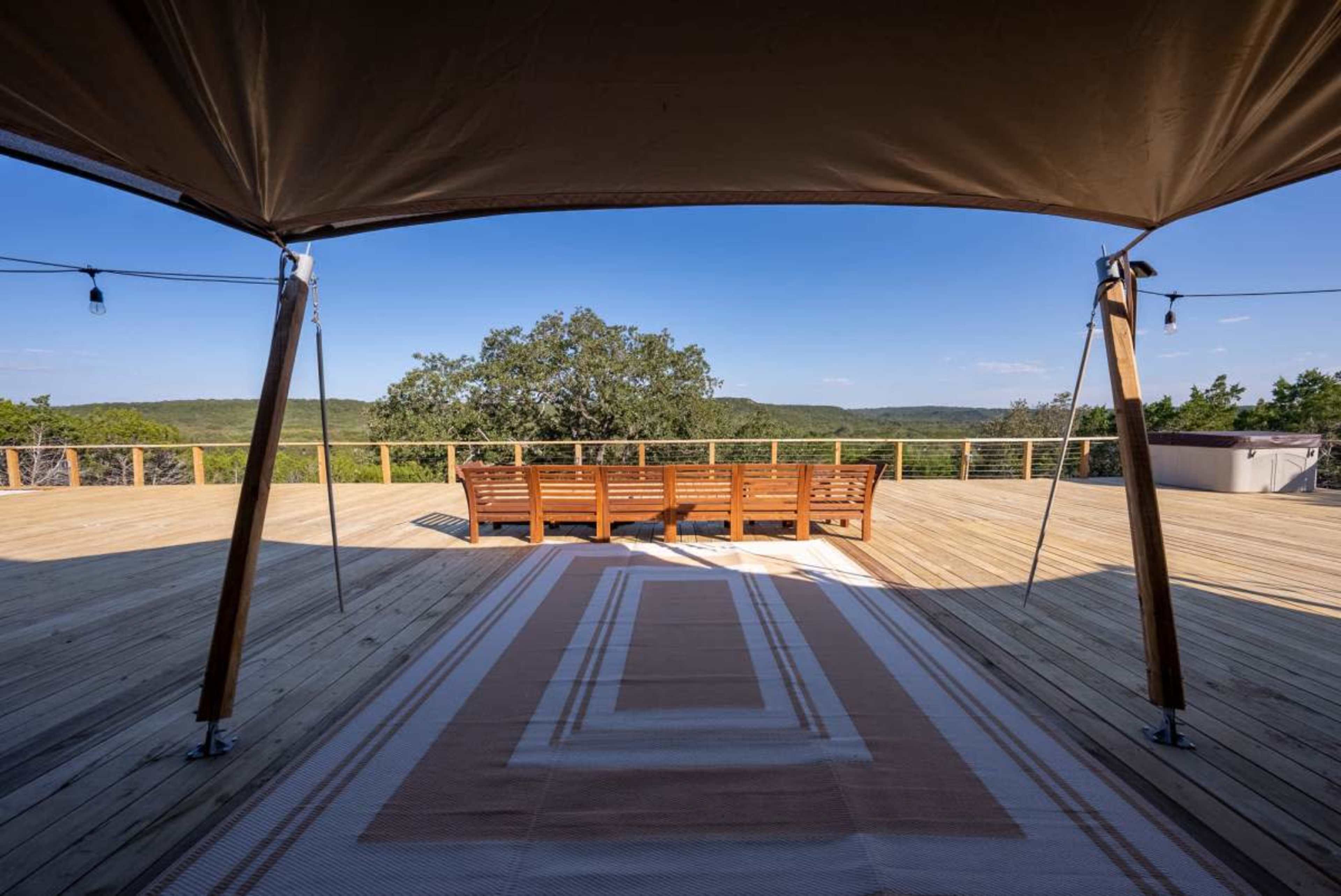 The image shows a spacious wooden deck with a canopy and a wooden seating arrangement overlooking a scenic landscape.