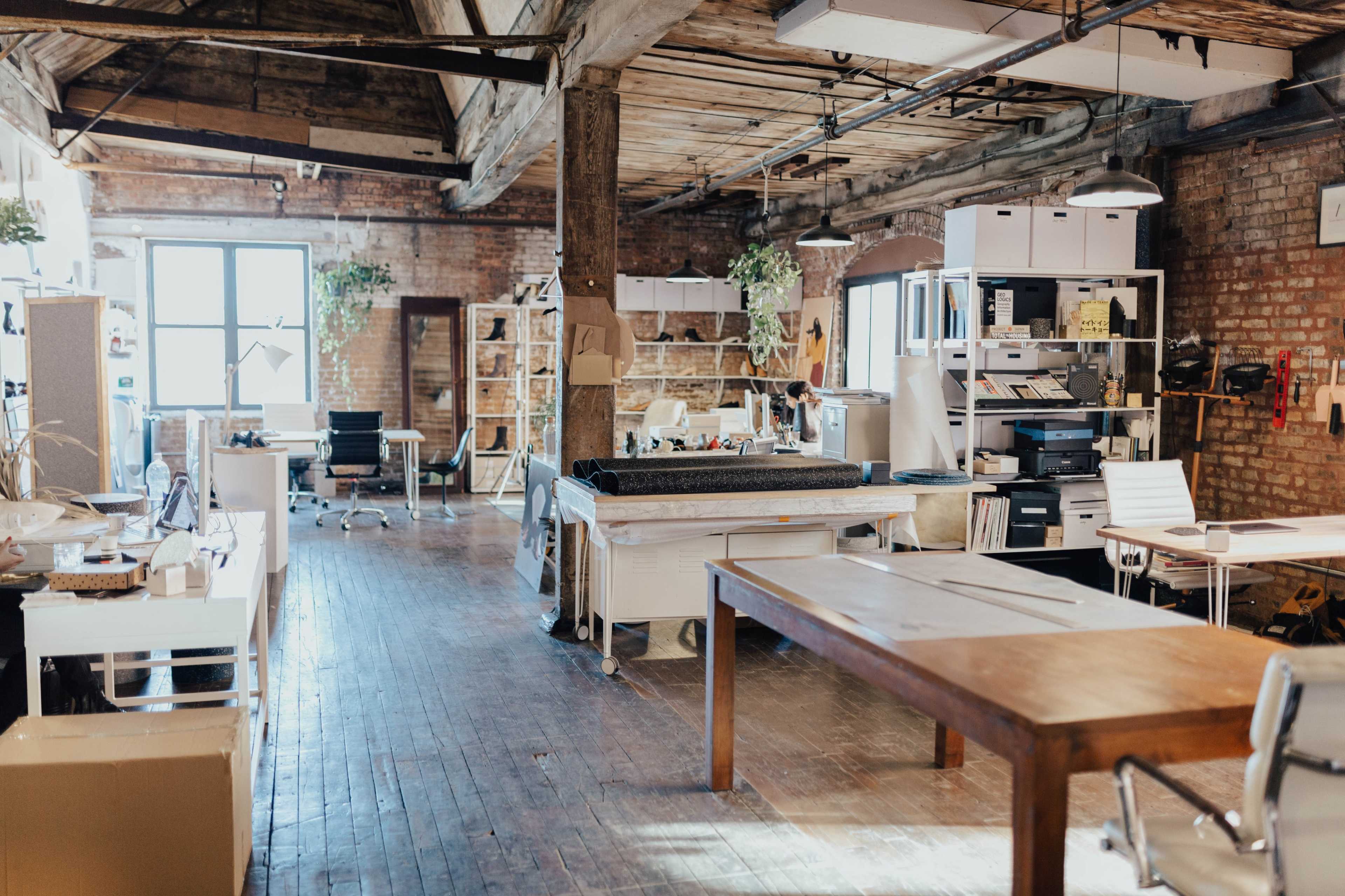 An open-plan office space featuring wooden beams, brick walls, multiple workstations, and a large table in the center.