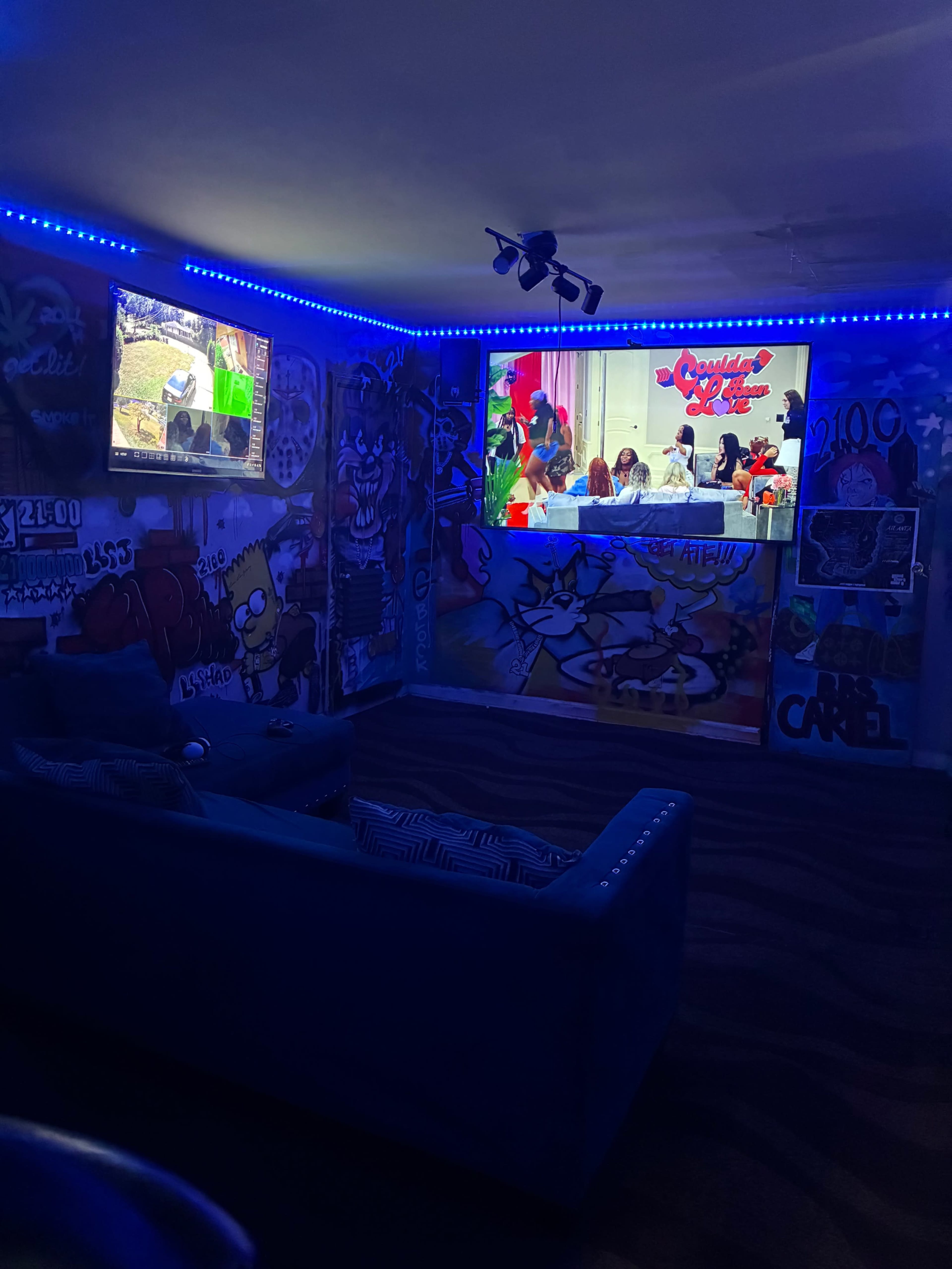 The image shows a themed room with bright blue LED lights, featuring a couch, a television displaying a video game, and a large wall screen showing a group of people gathered around a table, all surrounded by colorful graffiti art.