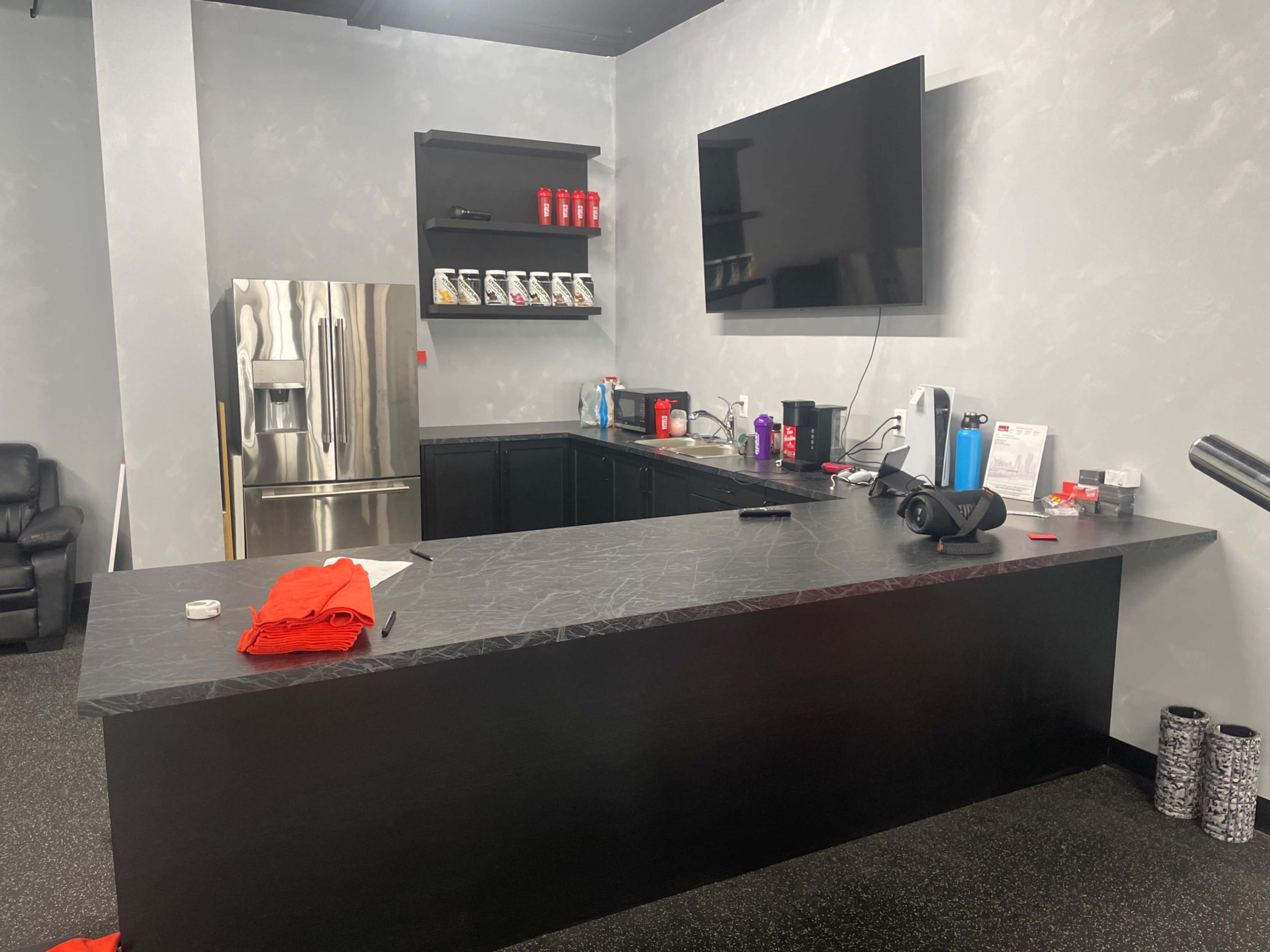 A modern, minimalist kitchenette features a large black countertop, a stainless steel refrigerator, and various appliances arranged on the counter.