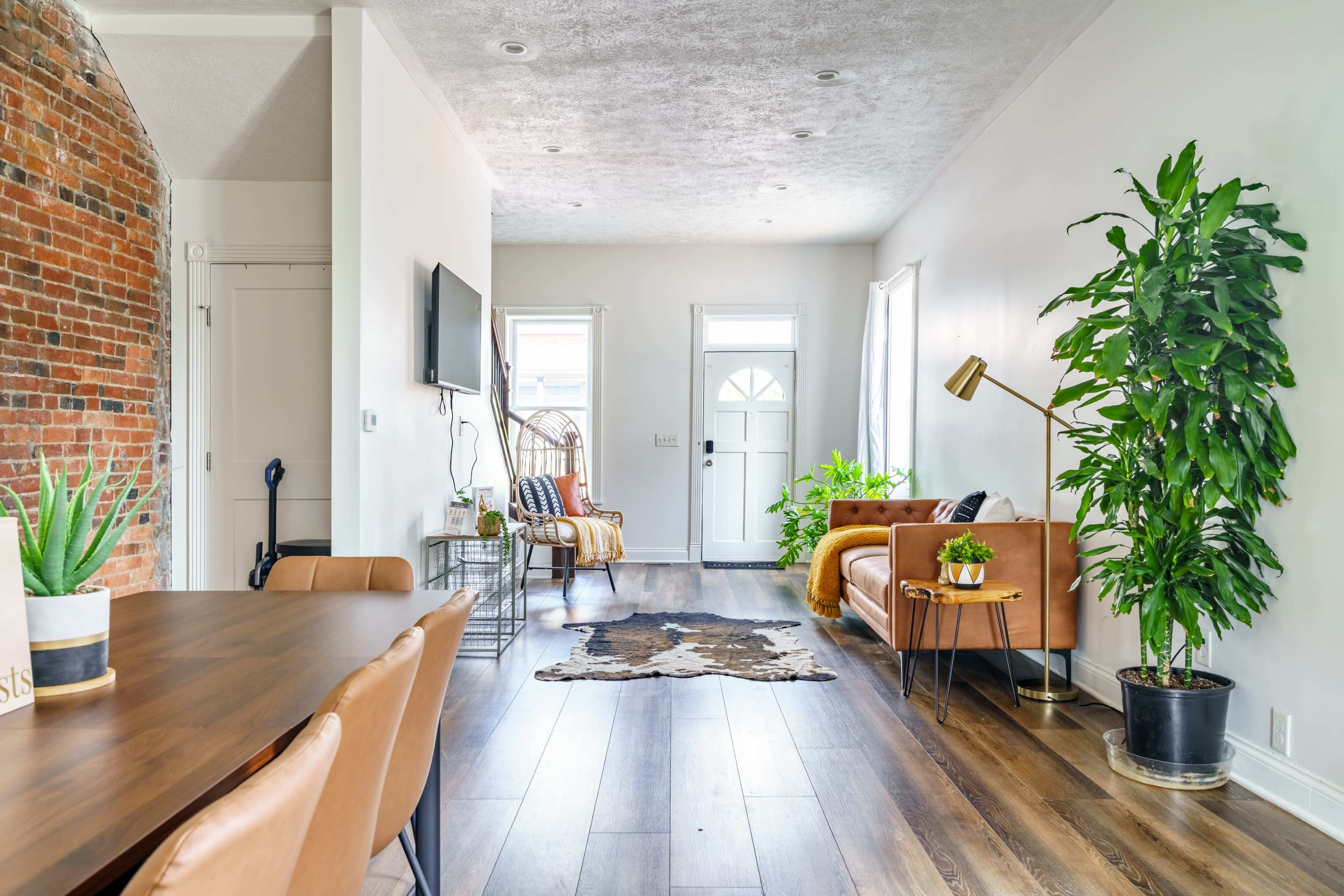The image shows a bright, modern living space featuring a brown leather sofa, a large potted plant, and a dining table, with exposed brick walls and large windows.