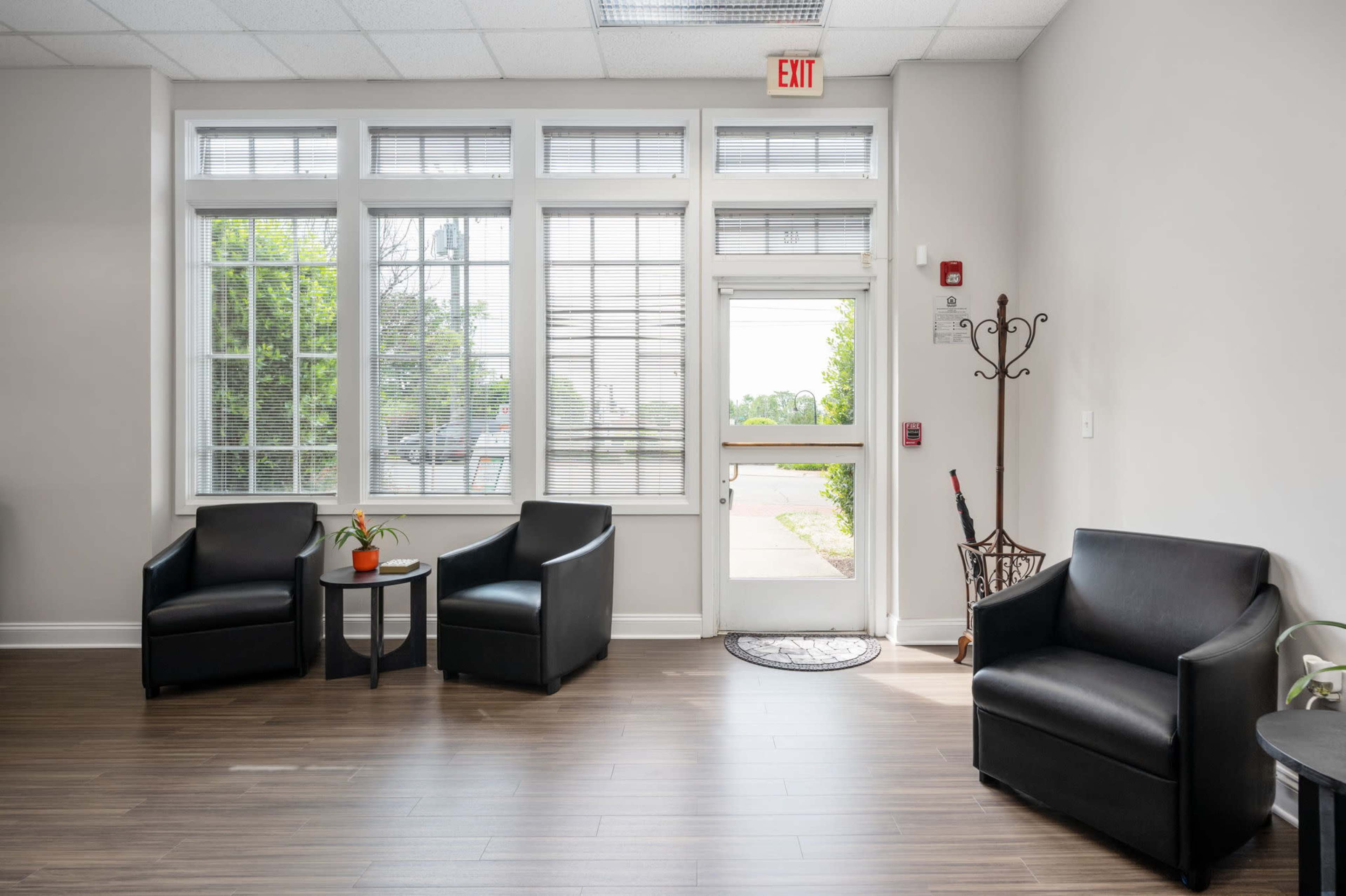 A waiting area with two black chairs, a small round table with a potted plant, and a door leading outside, surrounded by large windows.