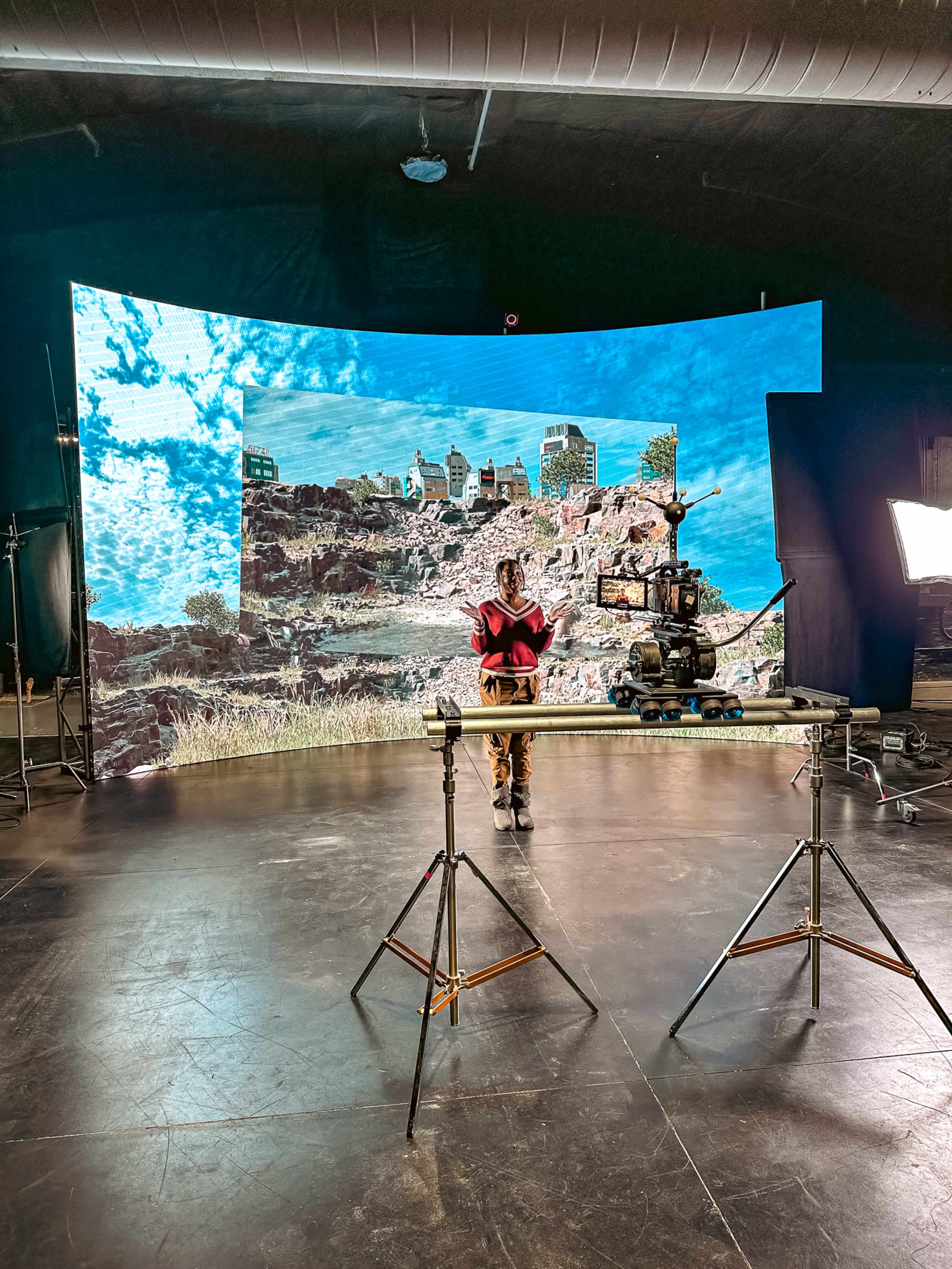 A person stands in front of a large curved screen displaying a landscape, with a camera setup on a tripod nearby in a studio environment.