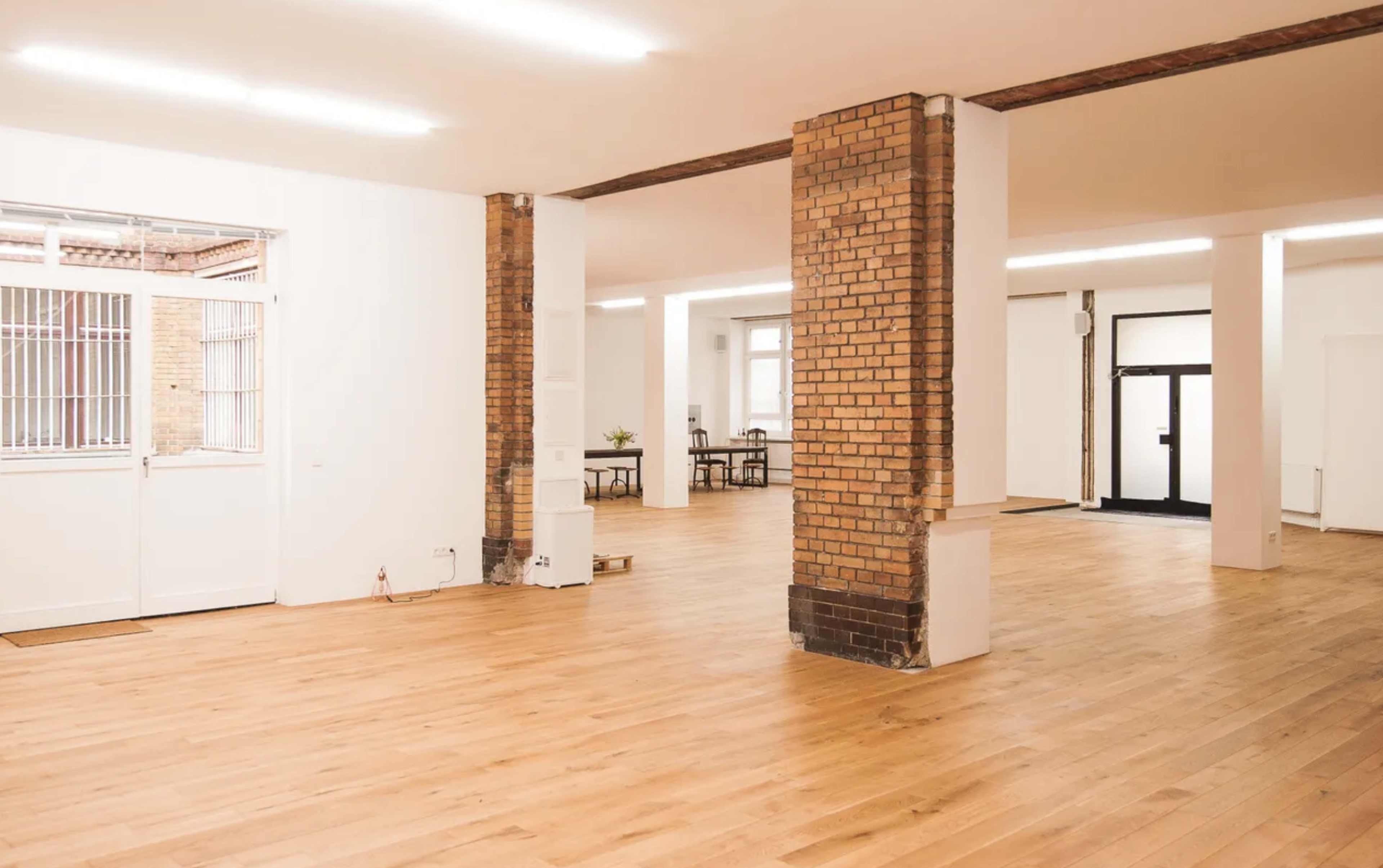 A spacious, empty room with wooden flooring, exposed brick columns, and large windows letting in natural light.