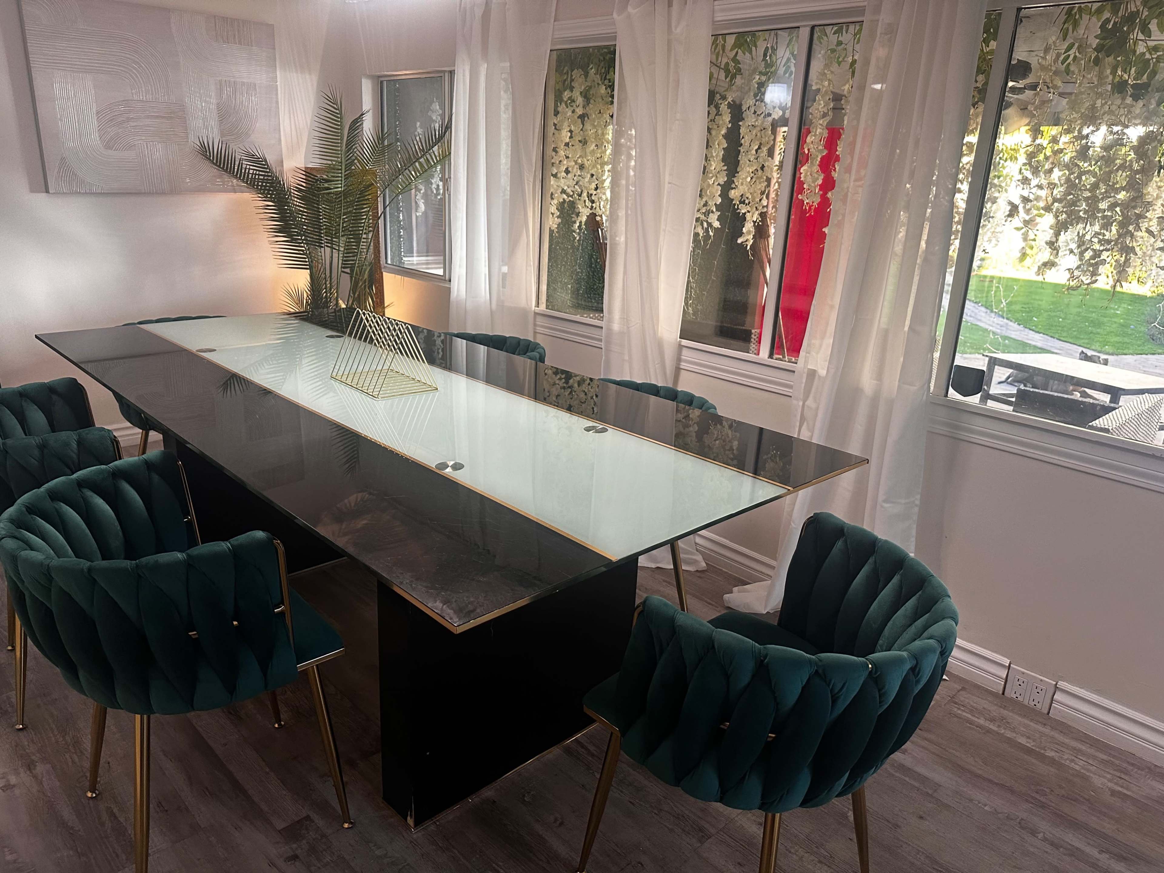 A modern conference room features a long glass table surrounded by green velvet chairs, with large windows letting in natural light.