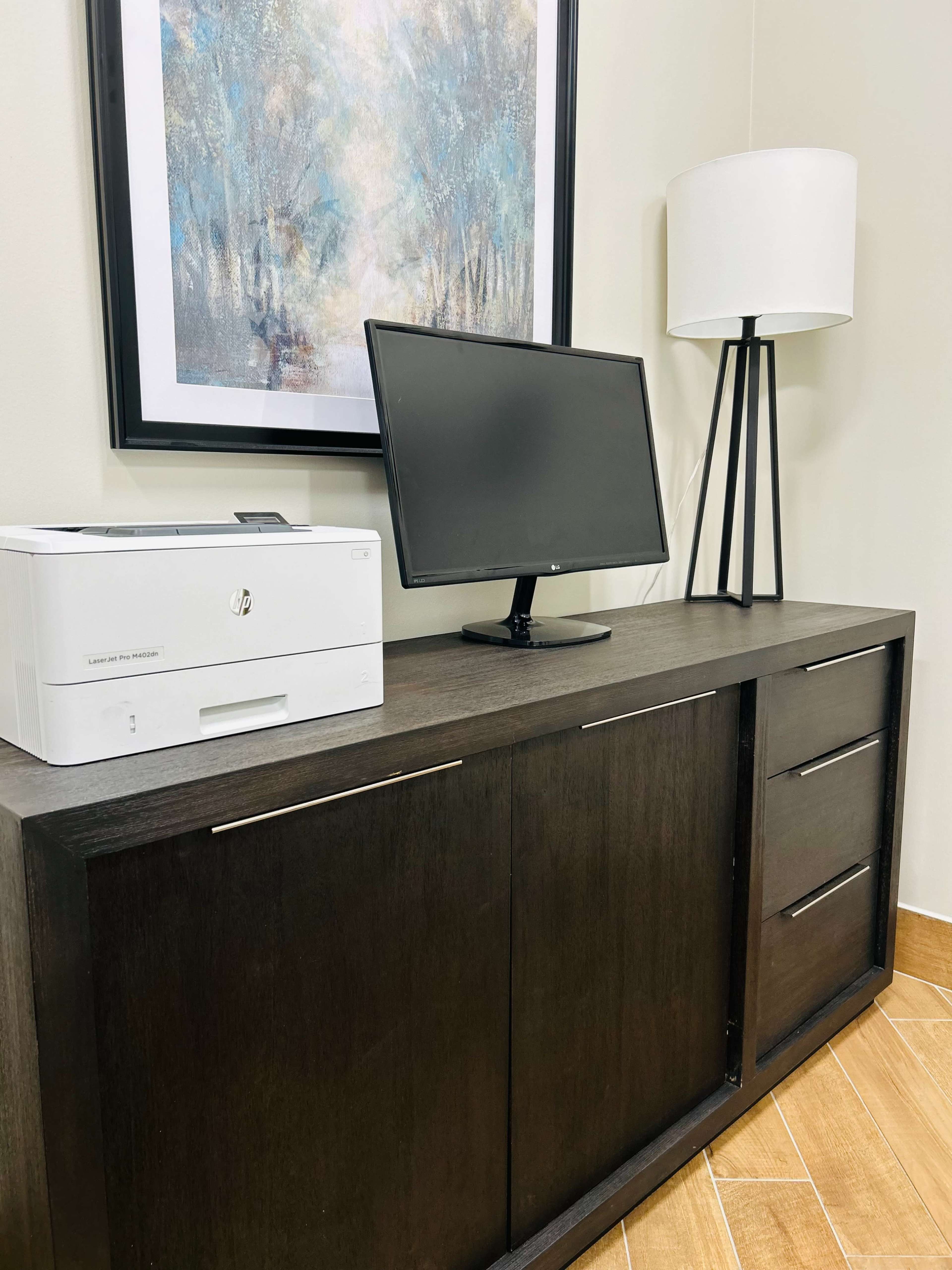 The image shows a modern office setup featuring a printer, a computer monitor on a stand, and a lamp, all placed on a dark wooden console with two drawers.