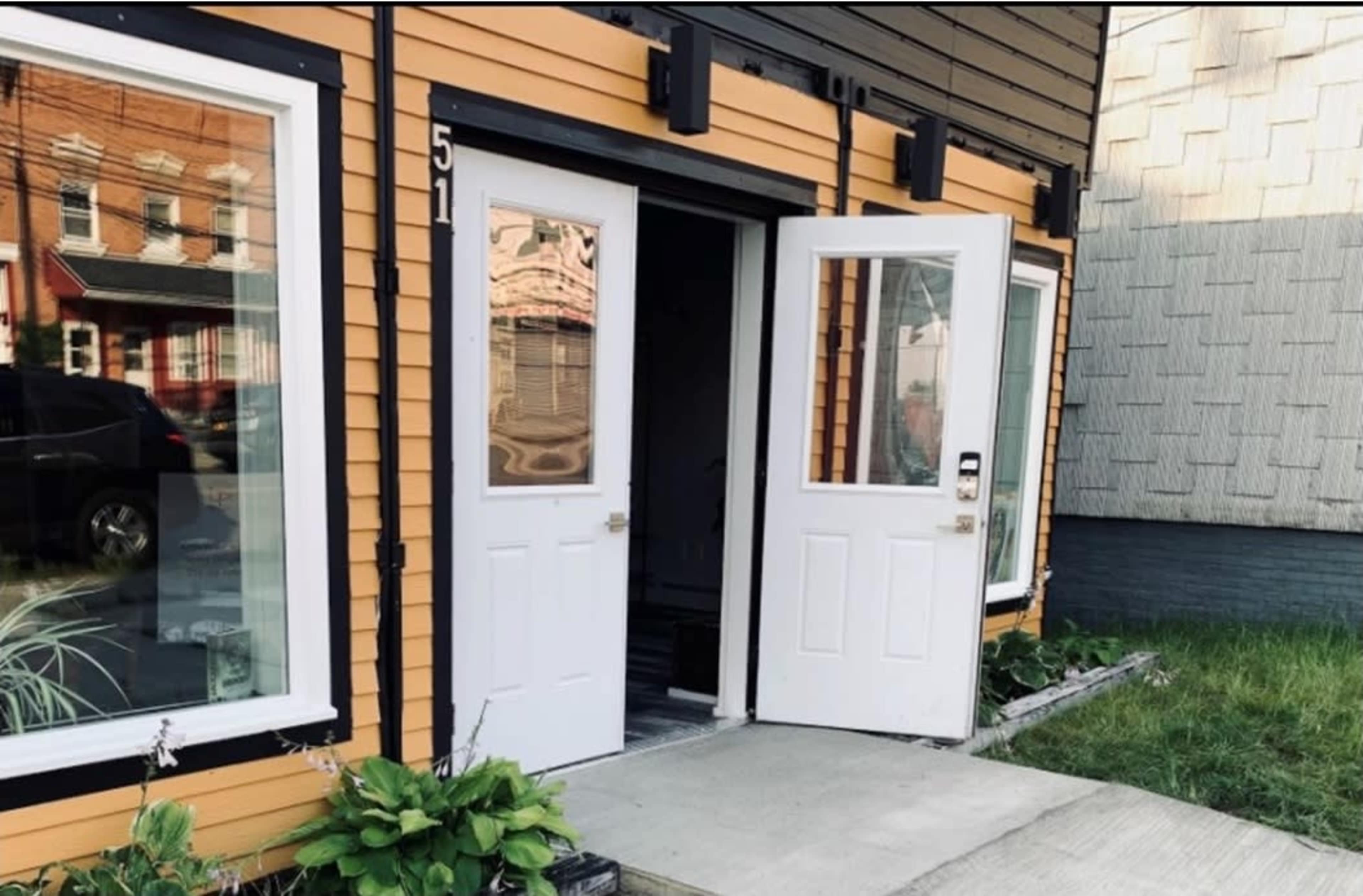 A building with orange and black siding, featuring two white doors, one of which is open, and a concrete pathway leading to the entrance.