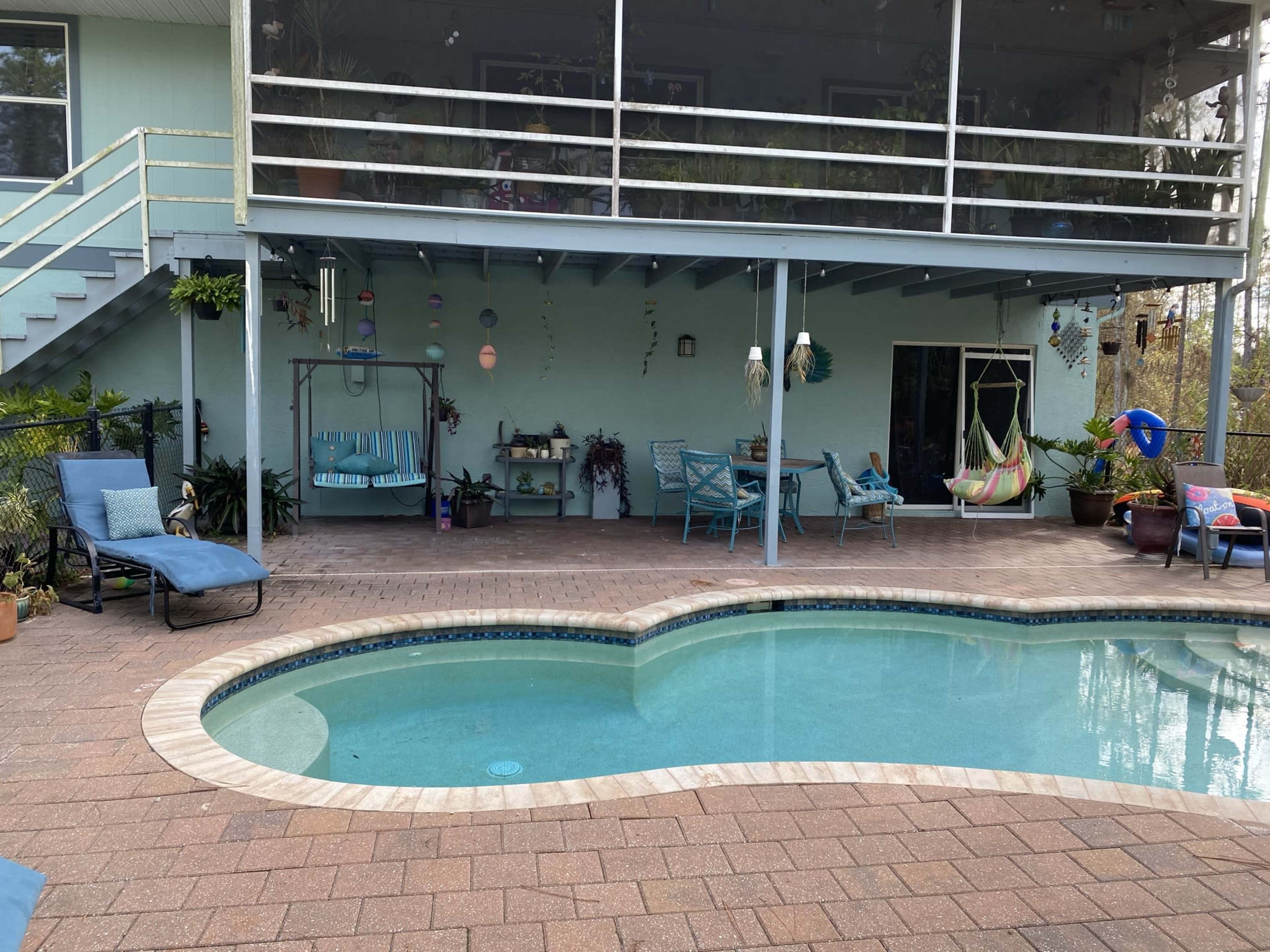 The image shows a swimming pool surrounded by a patio with seating areas, a covered deck above, and various plants and decorations.