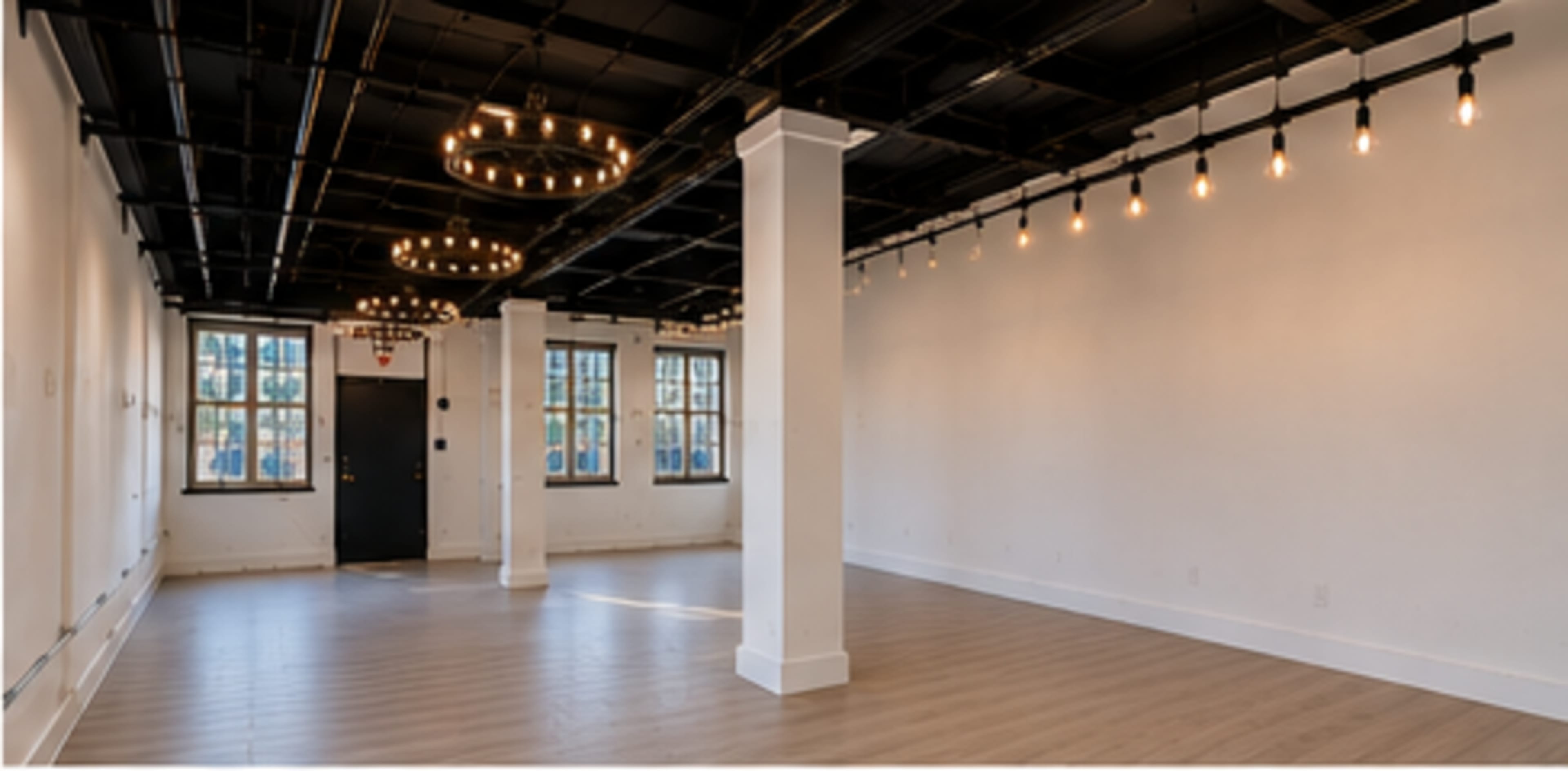 The image shows a spacious, empty room with white walls, wooden flooring, and several hanging light fixtures.