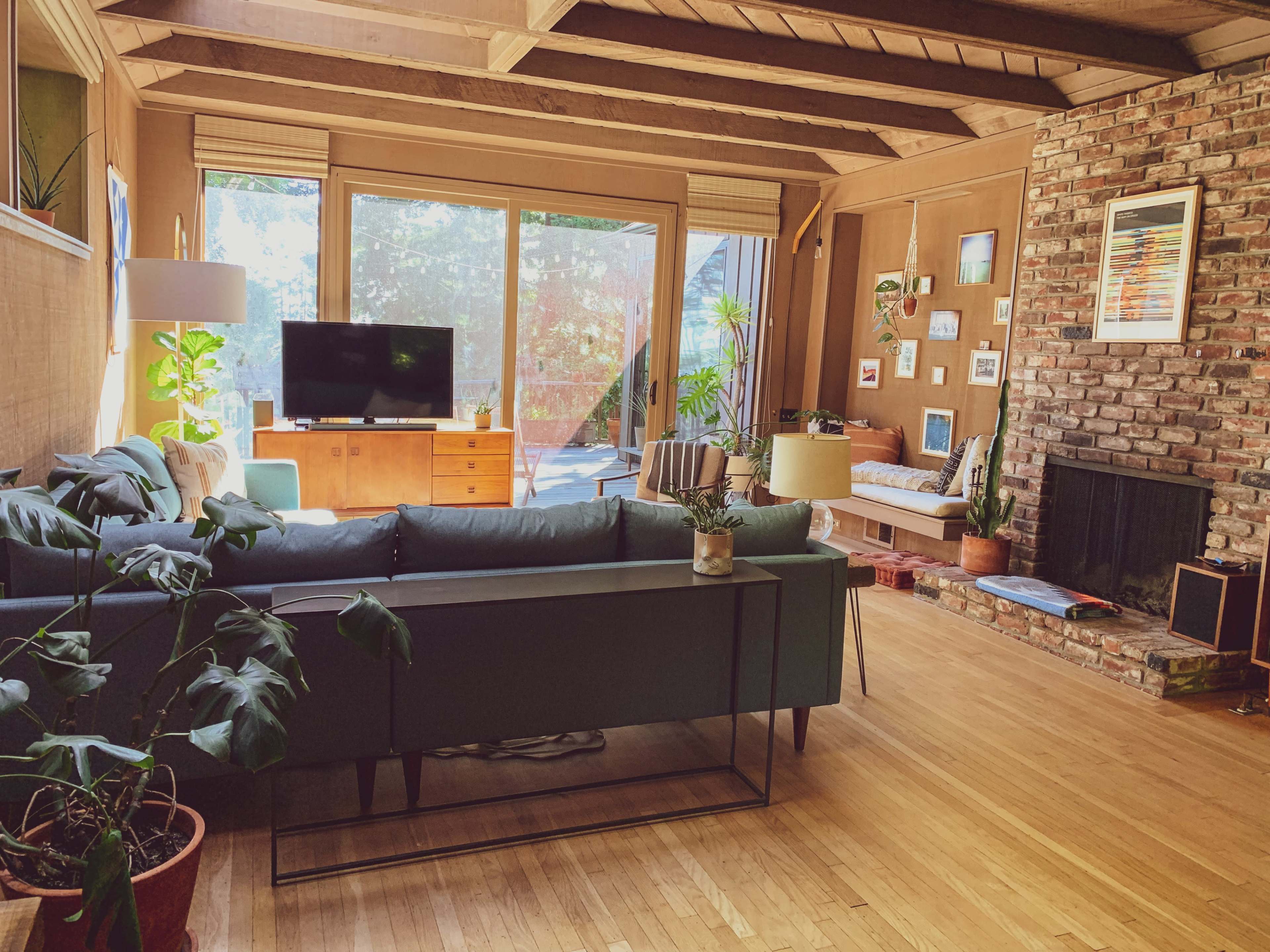 A cozy living room with a green sofa, a television on a wooden console, large windows allowing natural light, and a brick fireplace surrounded by various plants and wall art.
