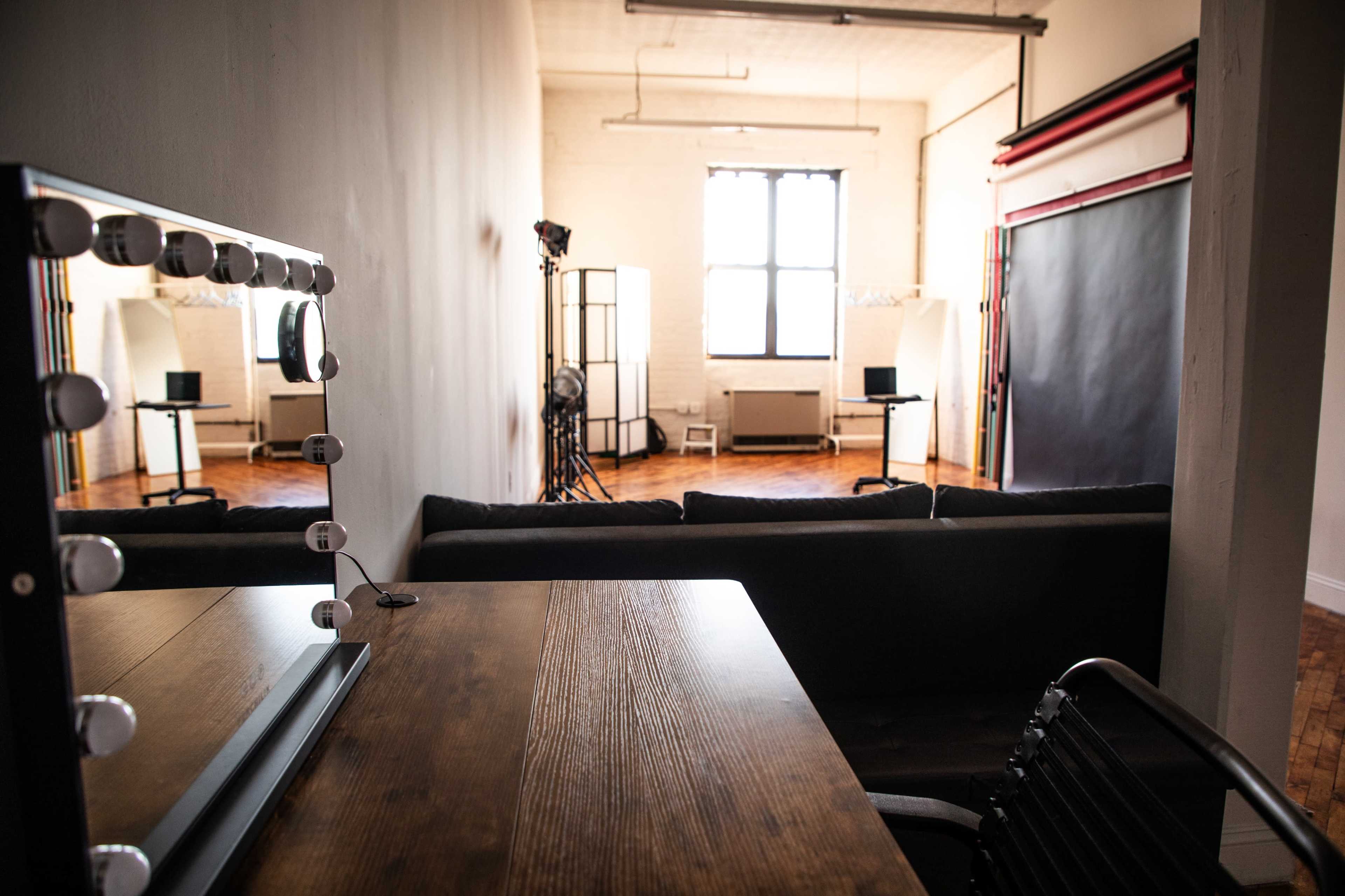 The image shows a spacious, well-lit studio featuring a vanity mirror with lights, a wooden desk, and a comfortable sofa, with photography equipment in the background.