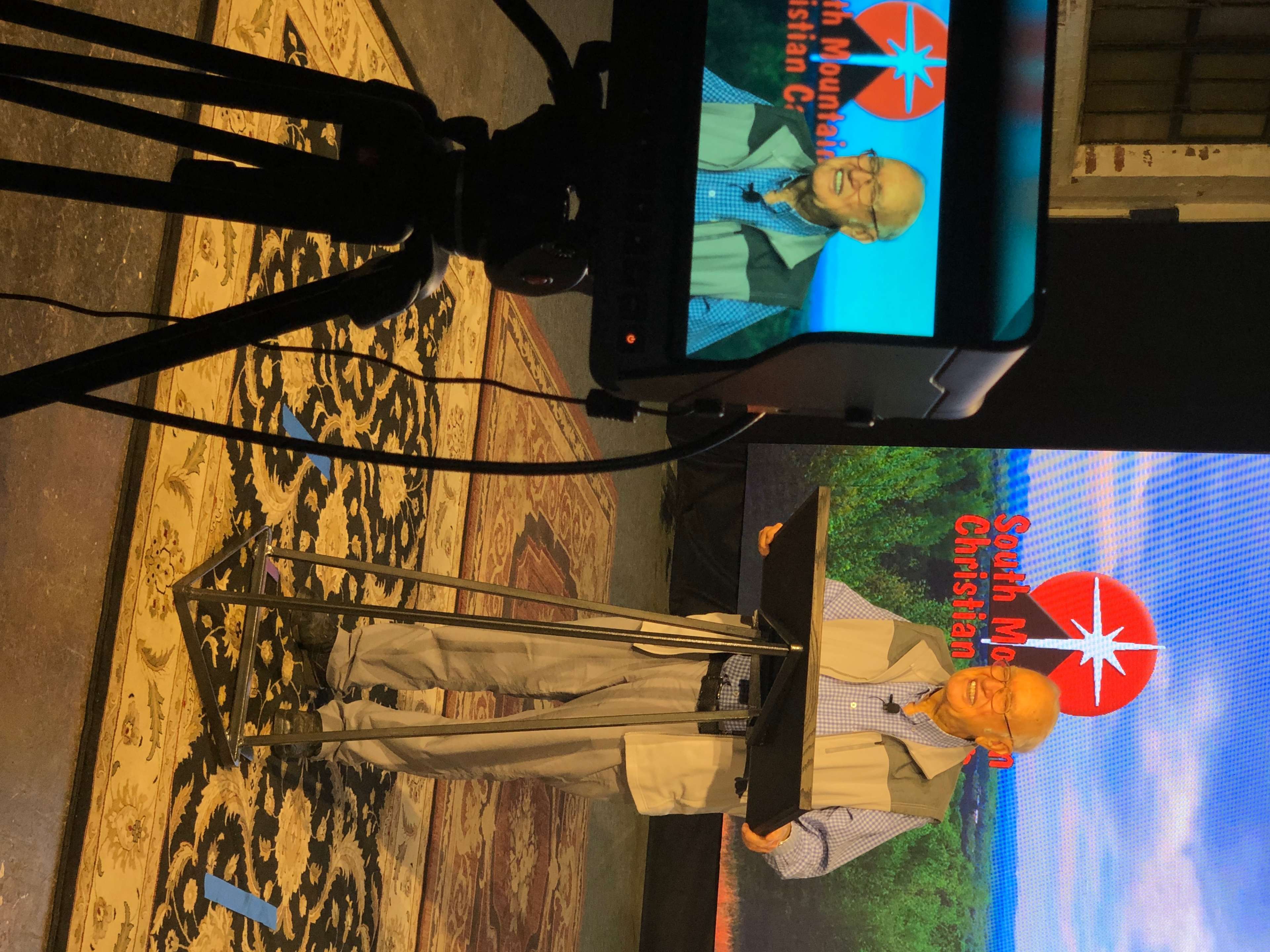 A speaker stands at a podium in front of a backdrop featuring the South Mountain Christian Center logo while addressing an audience through a camera setup.