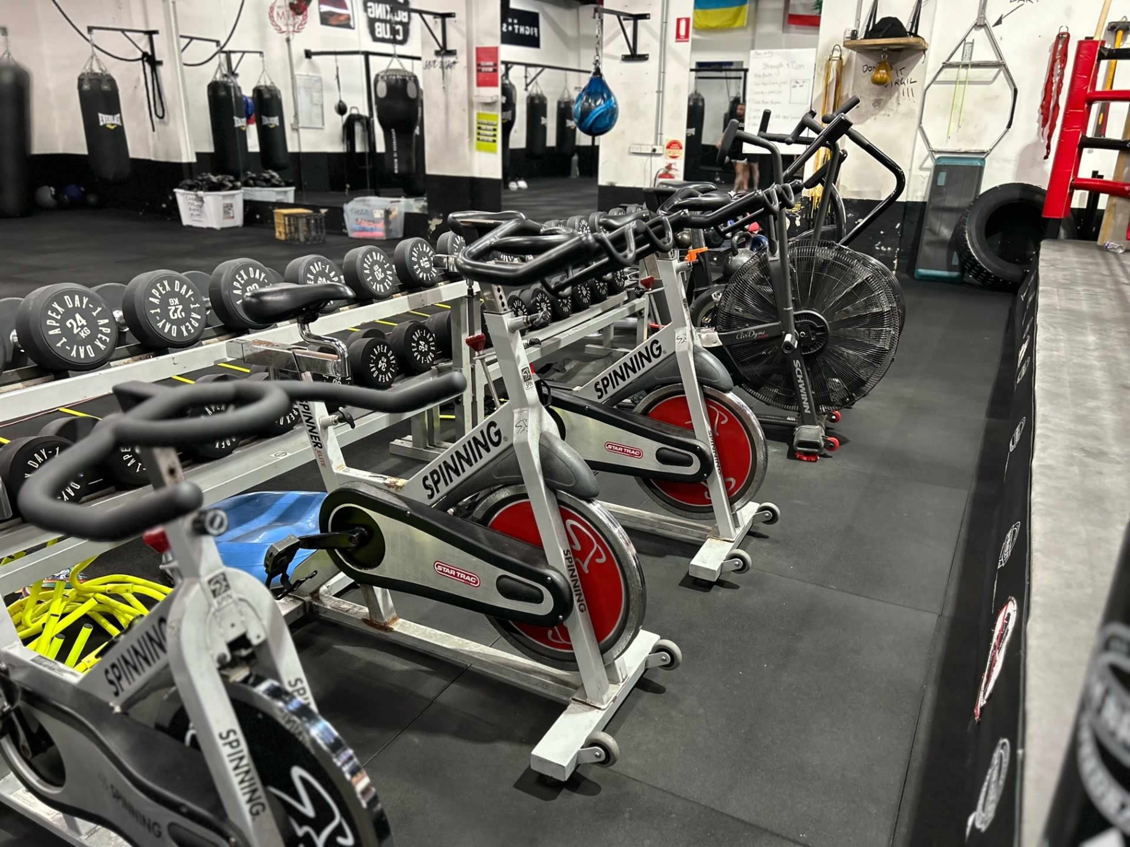 A row of stationary spinning bikes lined up against the wall in a gym, with various fitness equipment and punching bags in the background.