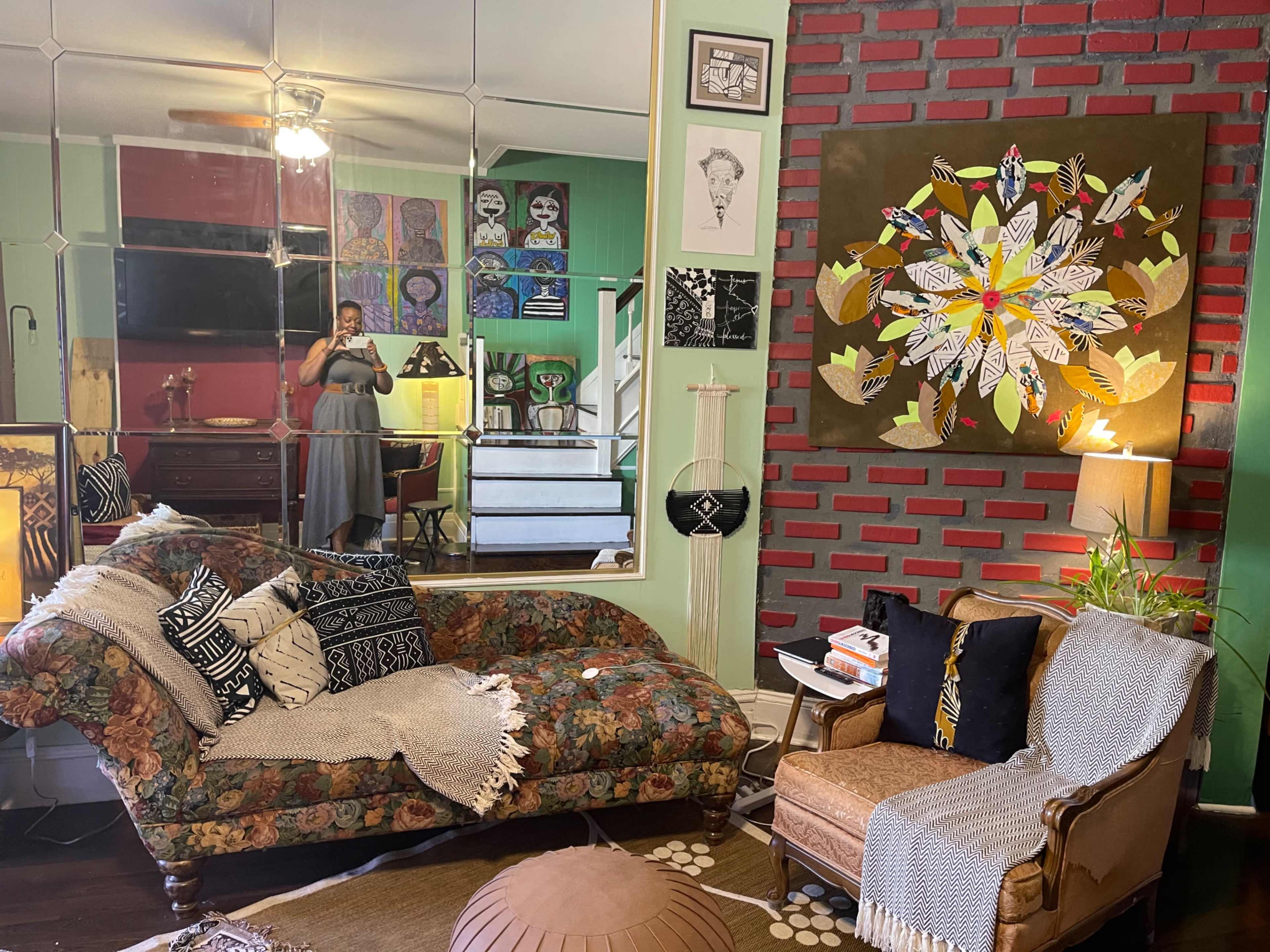 The room features a vintage floral sofa and a brown armchair, with a large wall adorned with colorful artwork and a mirror reflecting the space.