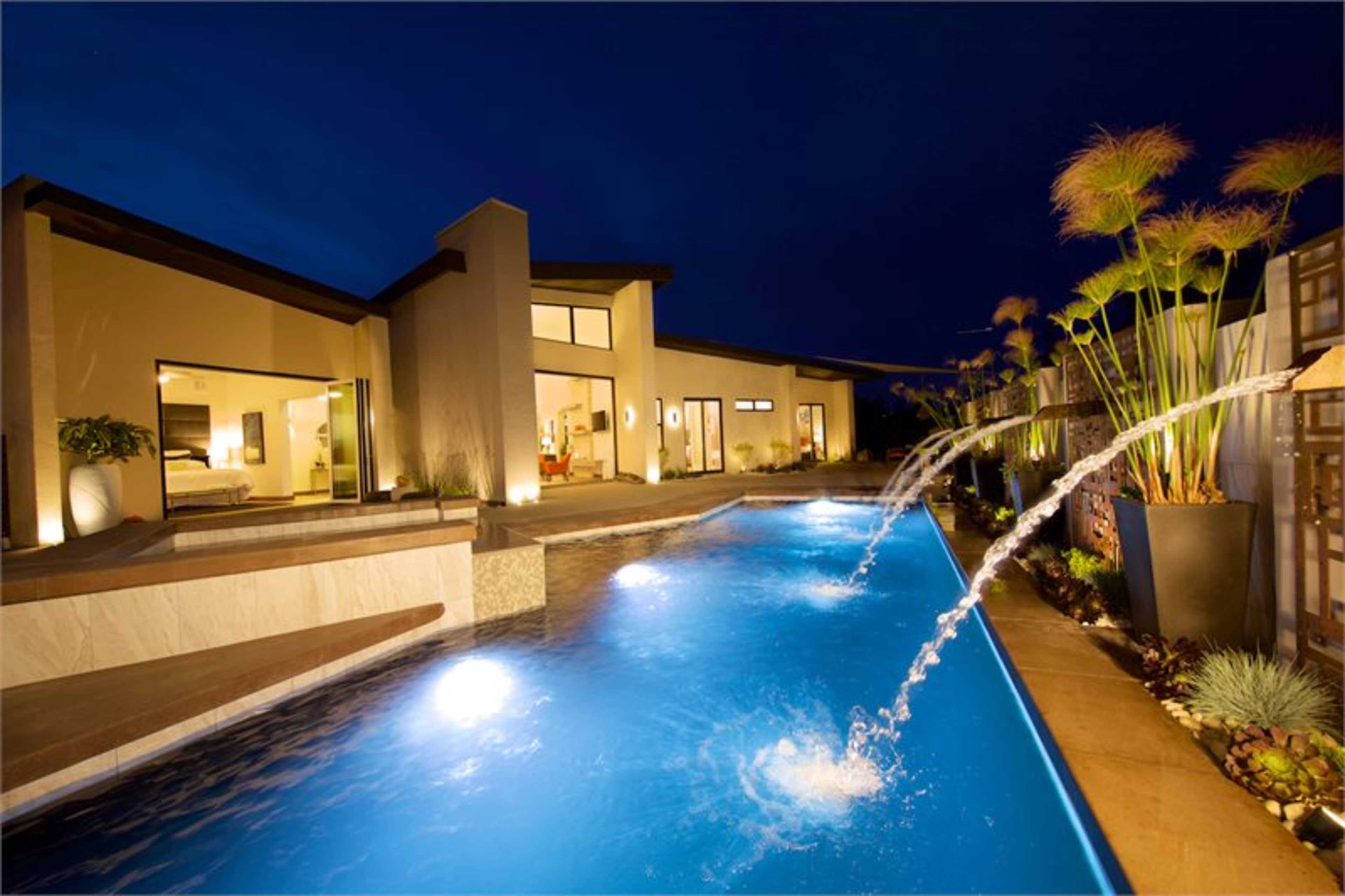 A modern home with large windows is illuminated at night, featuring a swimming pool with fountains and landscaping along the edges.