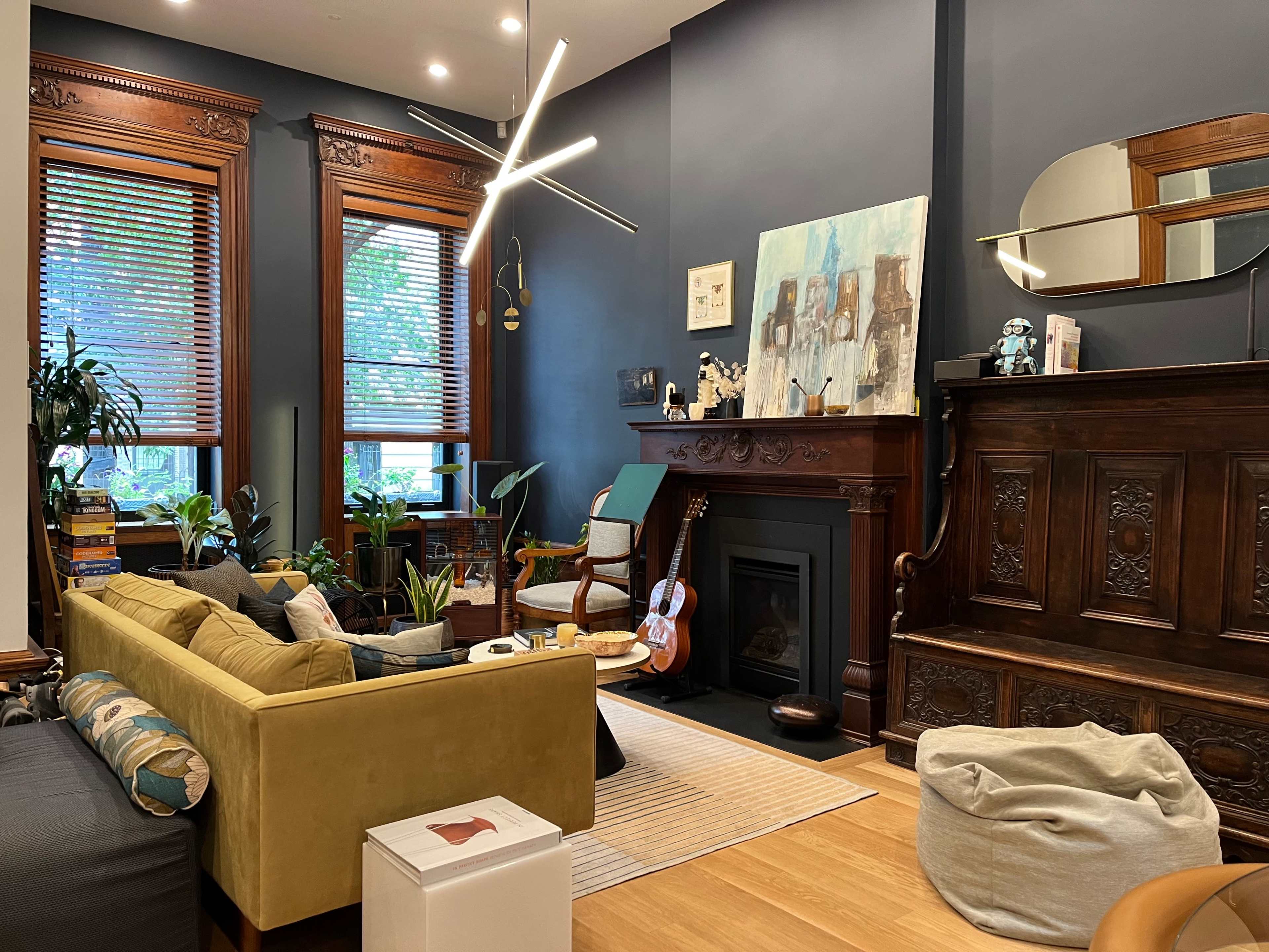 The living room features a green sofa, a guitar, plants, and a decorative wooden fireplace with a wall art piece above it.
