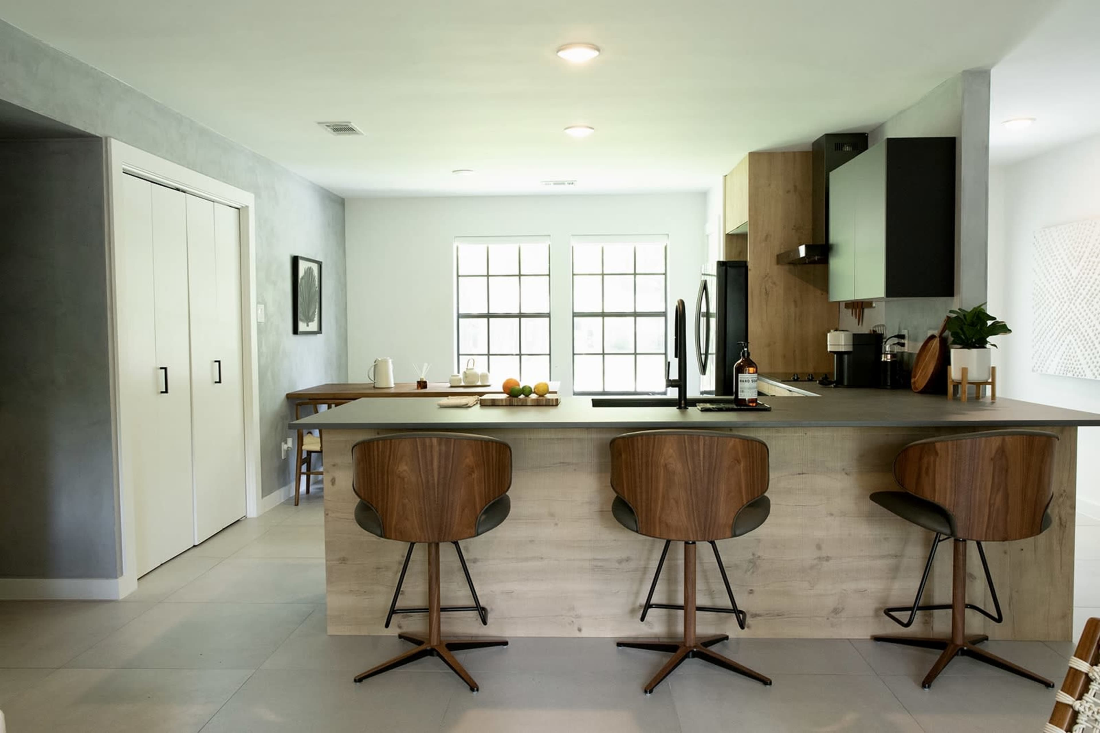 A modern kitchen features a large island with three bar stools, a dining table, and large windows that provide natural light.