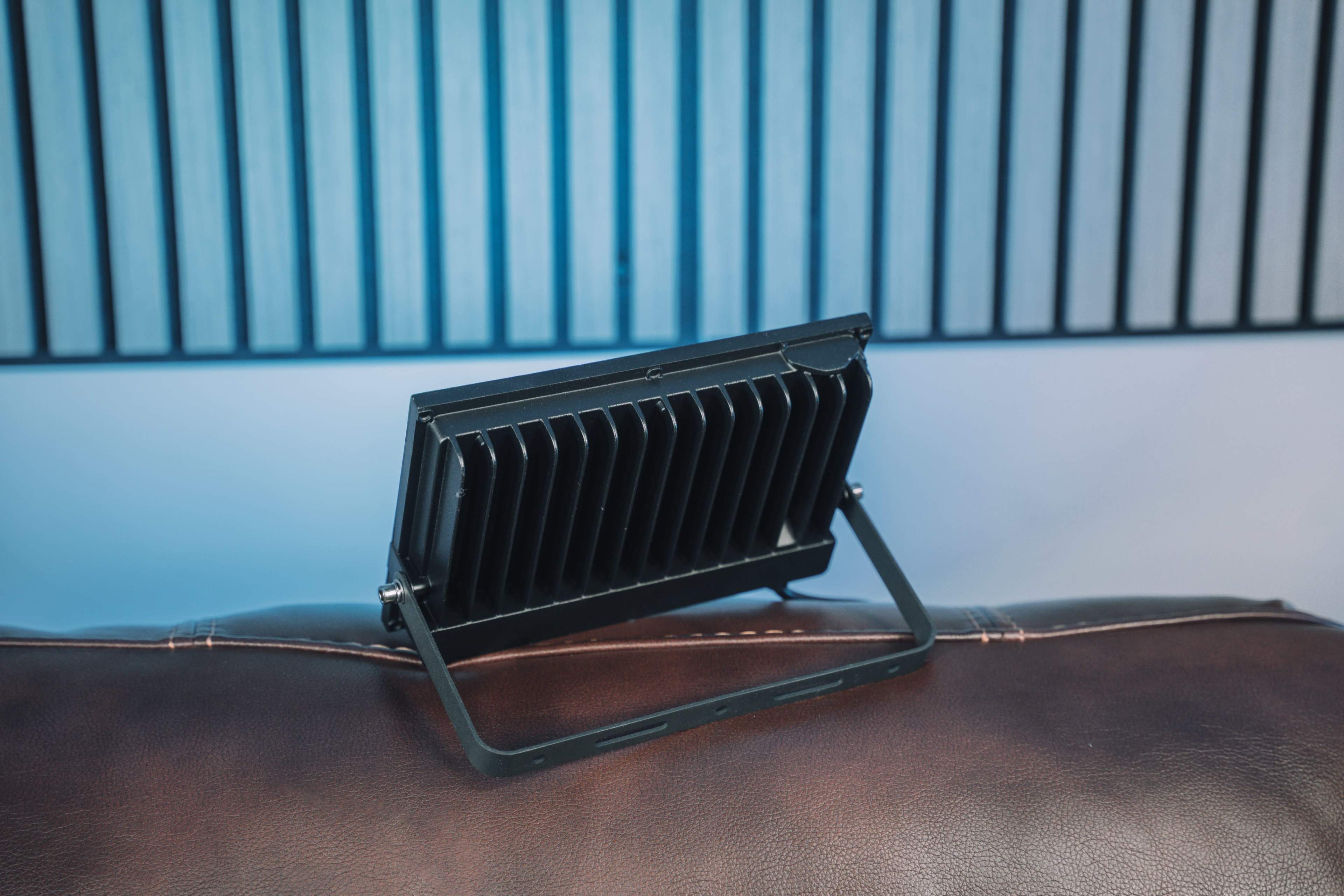 The image shows a black portable fan with a stand positioned on a brown leather surface, set against a backdrop of horizontal acoustic panels.