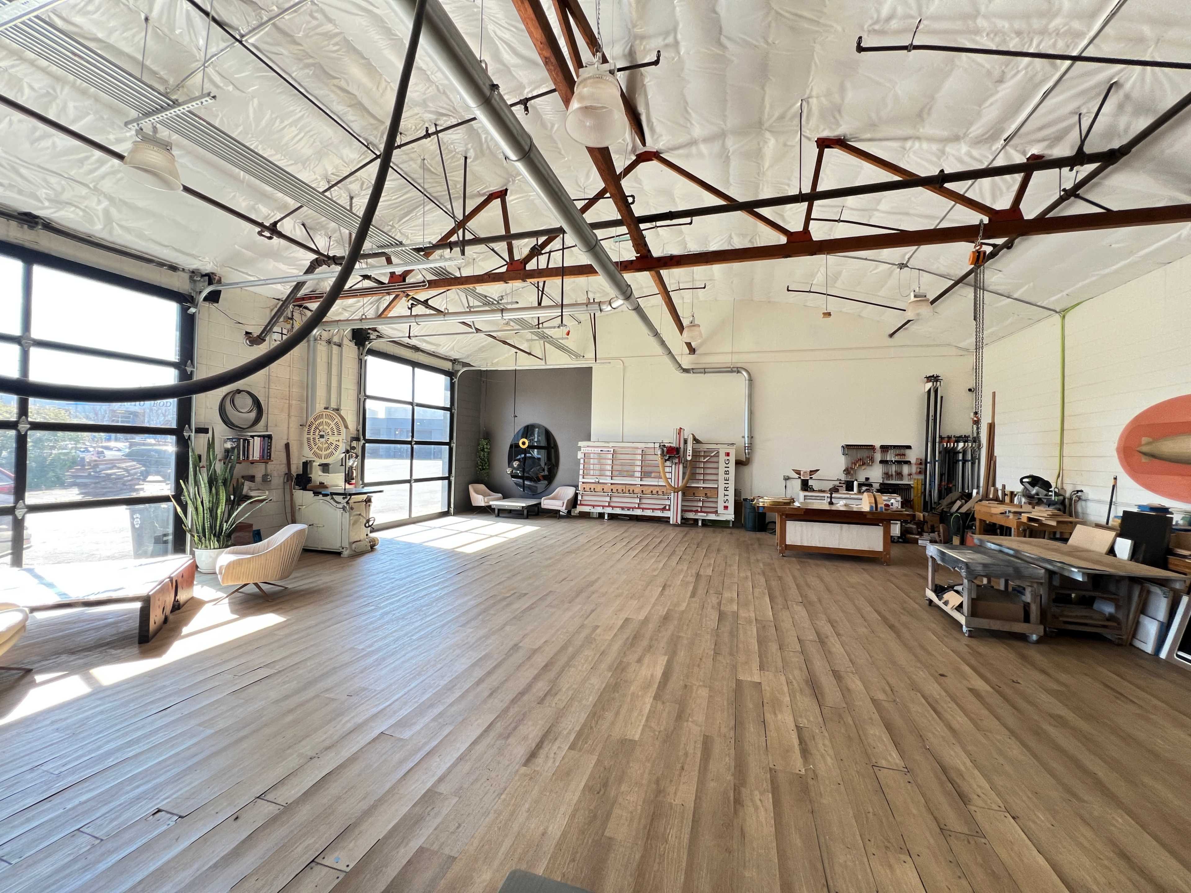 The image shows a spacious, well-lit interior of a studio or workshop with wooden flooring and large windows.