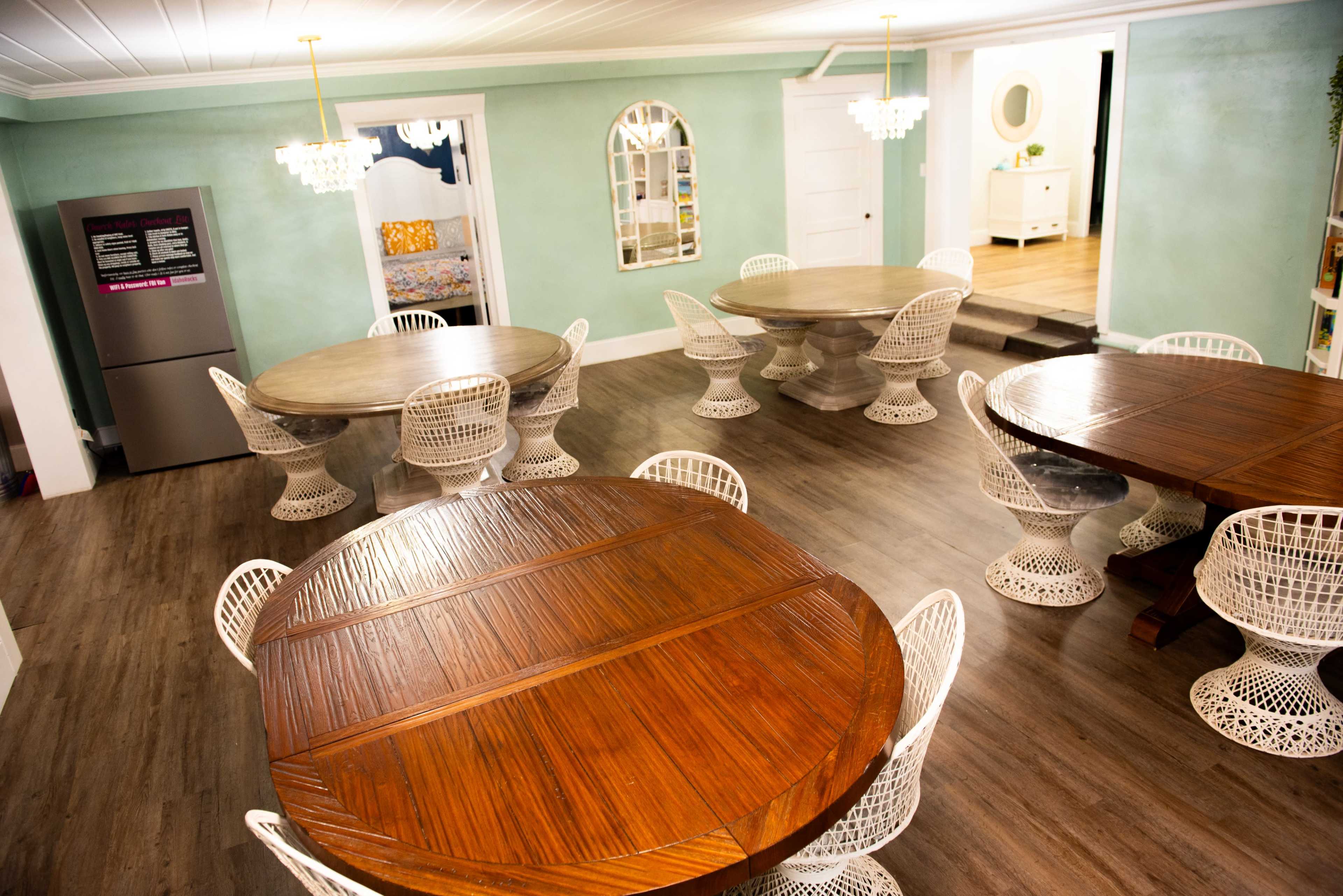 The image shows a well-lit dining area featuring multiple round wooden tables surrounded by white woven chairs, with green walls and a modern refrigerator in the background.