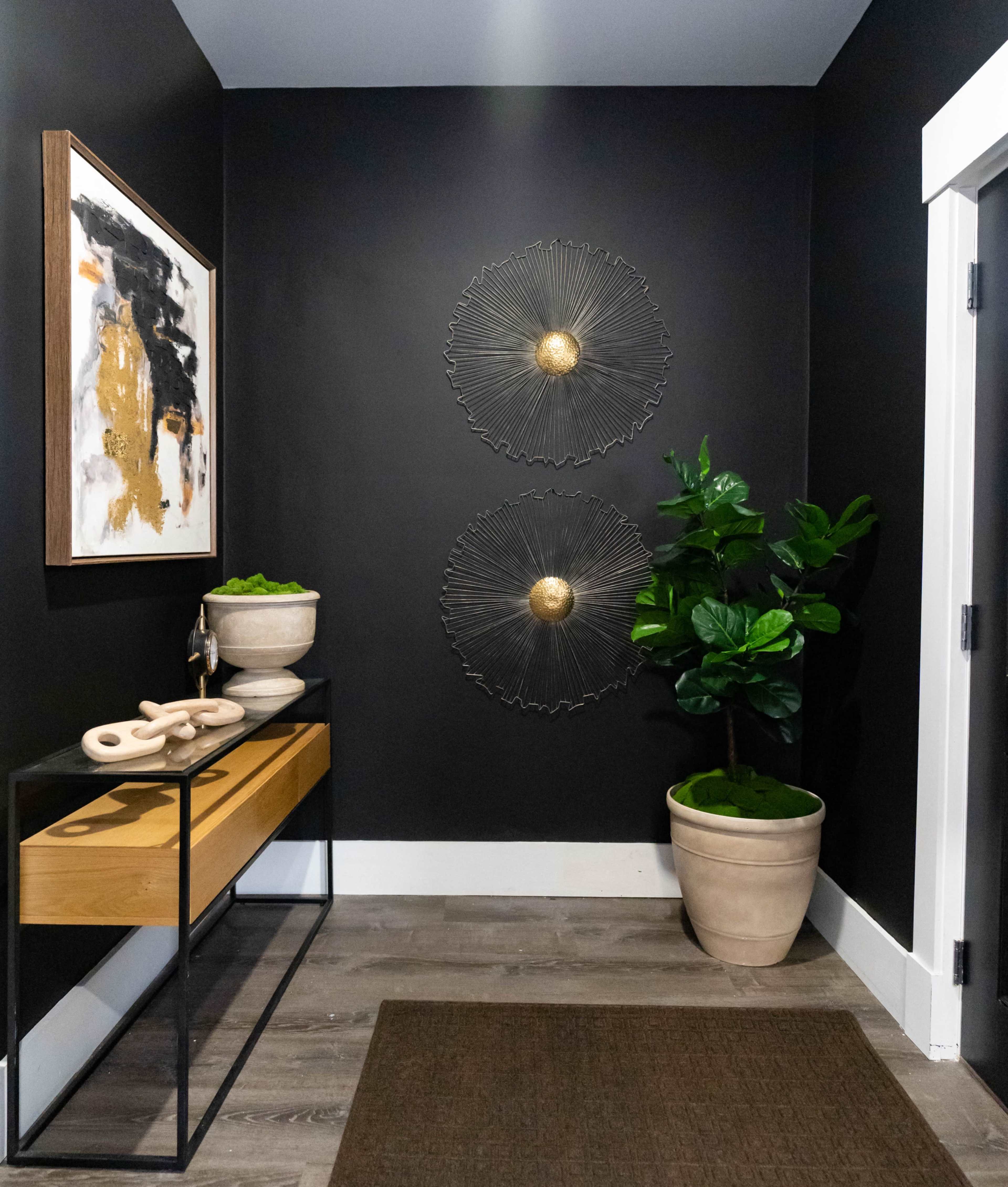 A modern entryway features dark walls, a wooden console table, two decorative wall hangings, and potted plants.