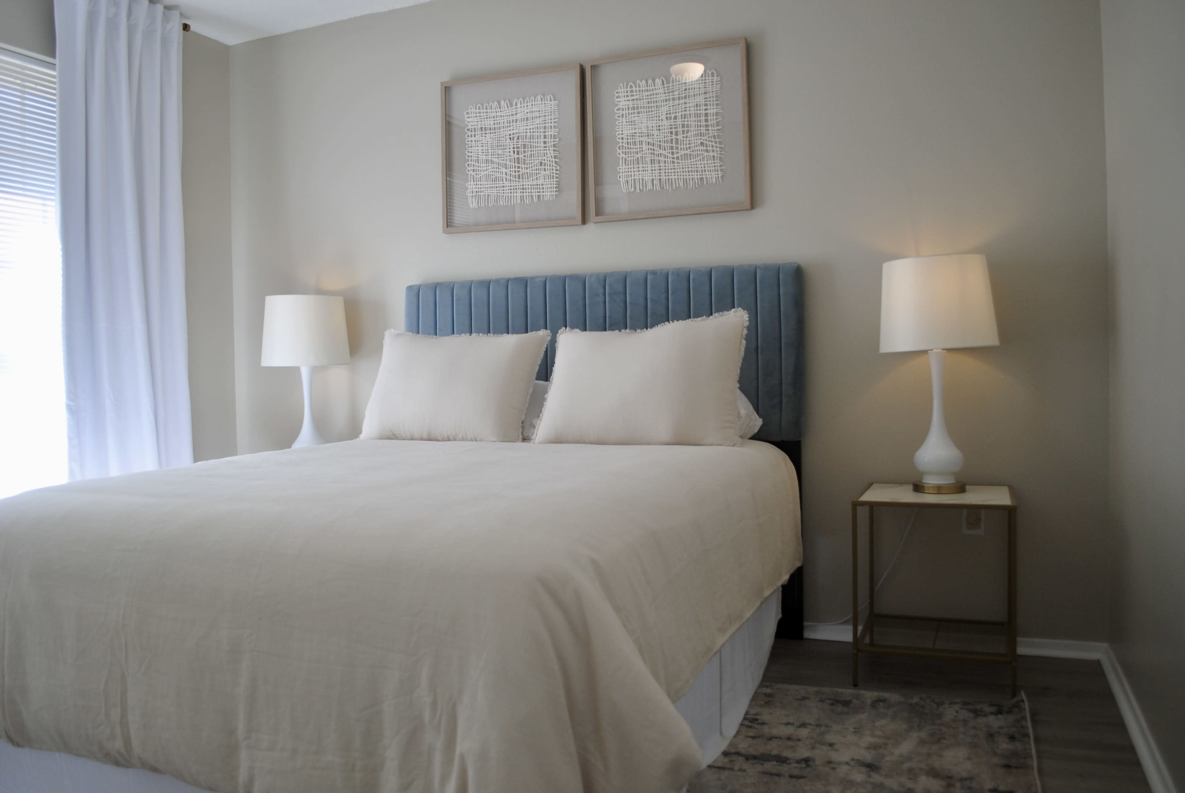 A neatly made bed with white linens and two pillows, positioned against a blue upholstered headboard in a softly lit room with framed artwork and two lamps on nightstands.