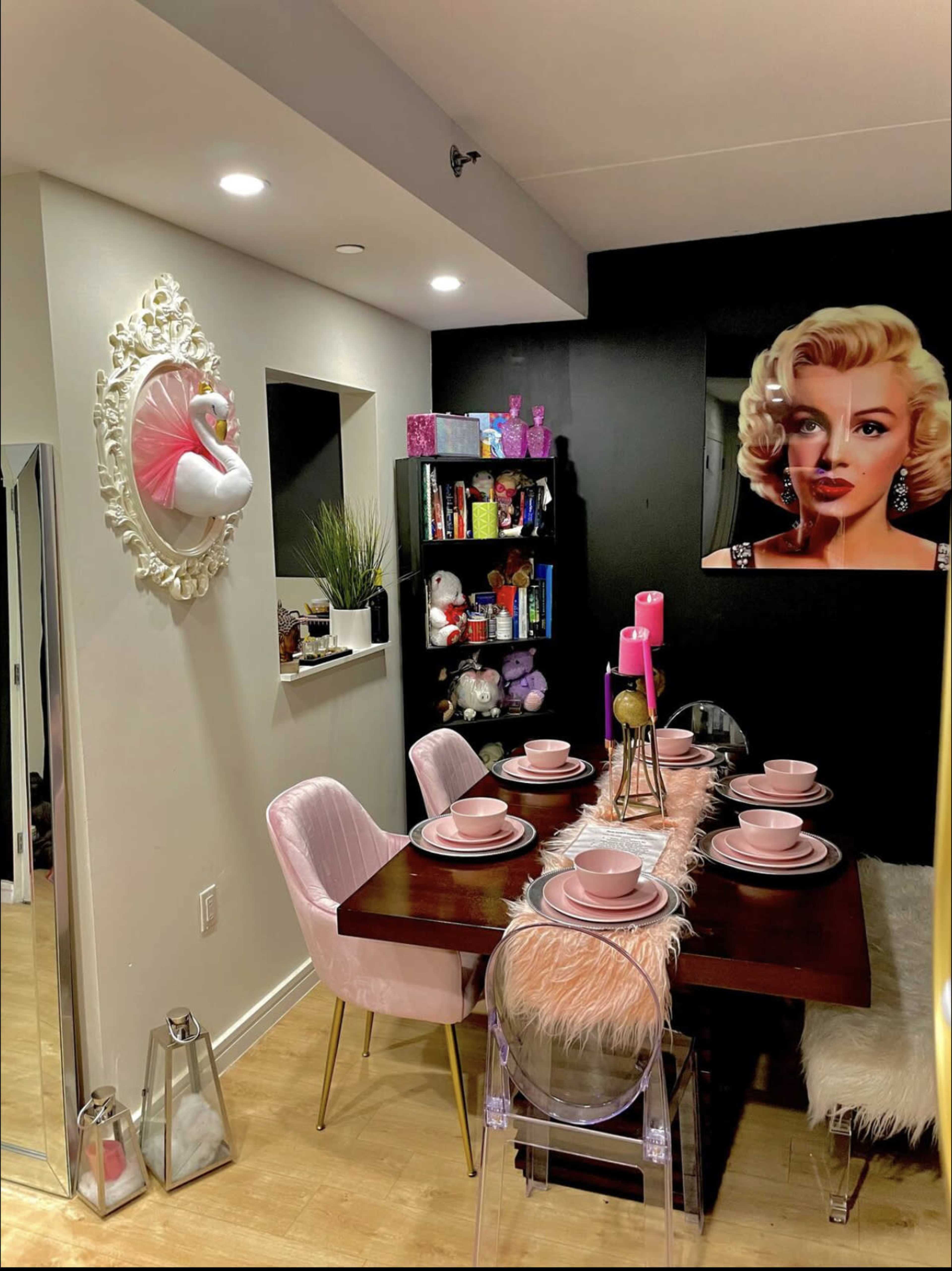 A dining area features a dark wall with a large portrait, a table set with pink dishes, and decorative items on shelves and surfaces.