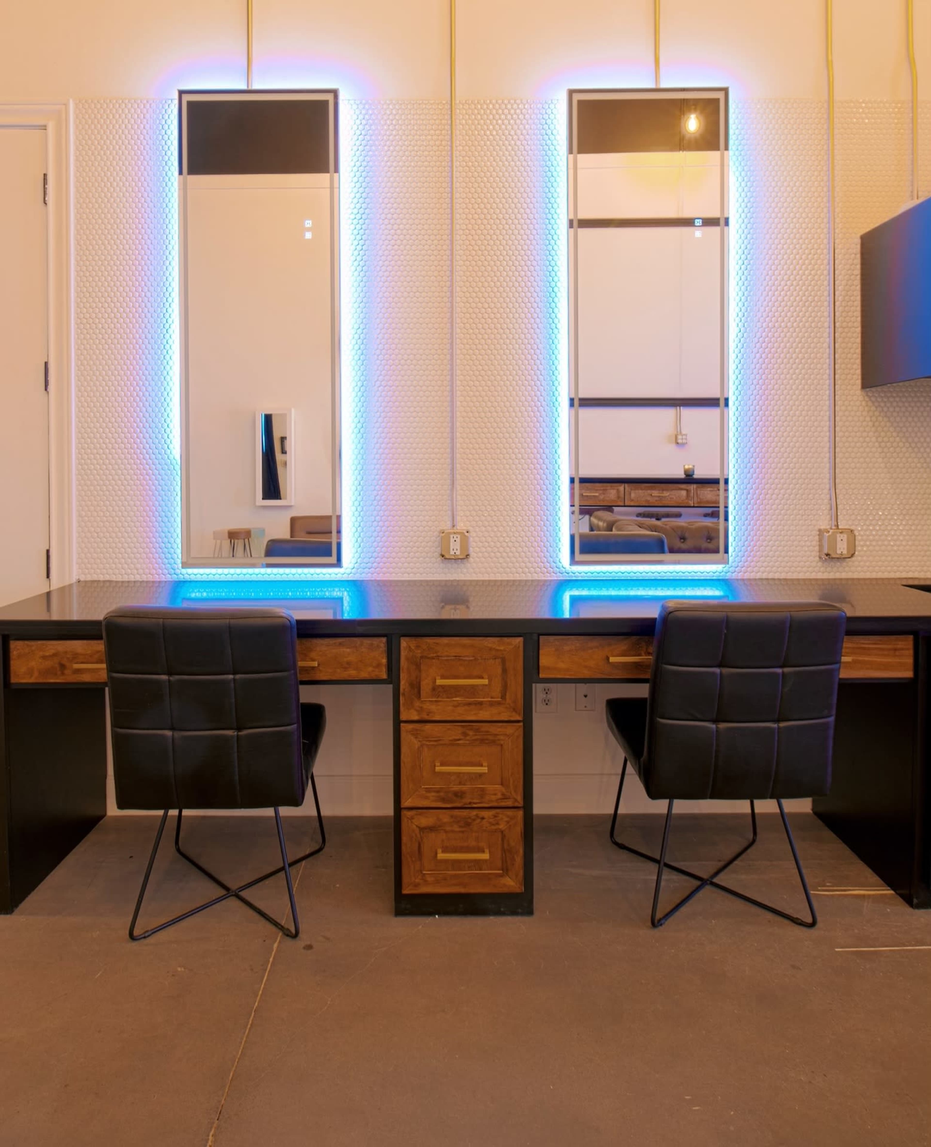 A modern vanity area with two illuminated mirrors above a sleek black desk and two black chairs, set against a textured wall.