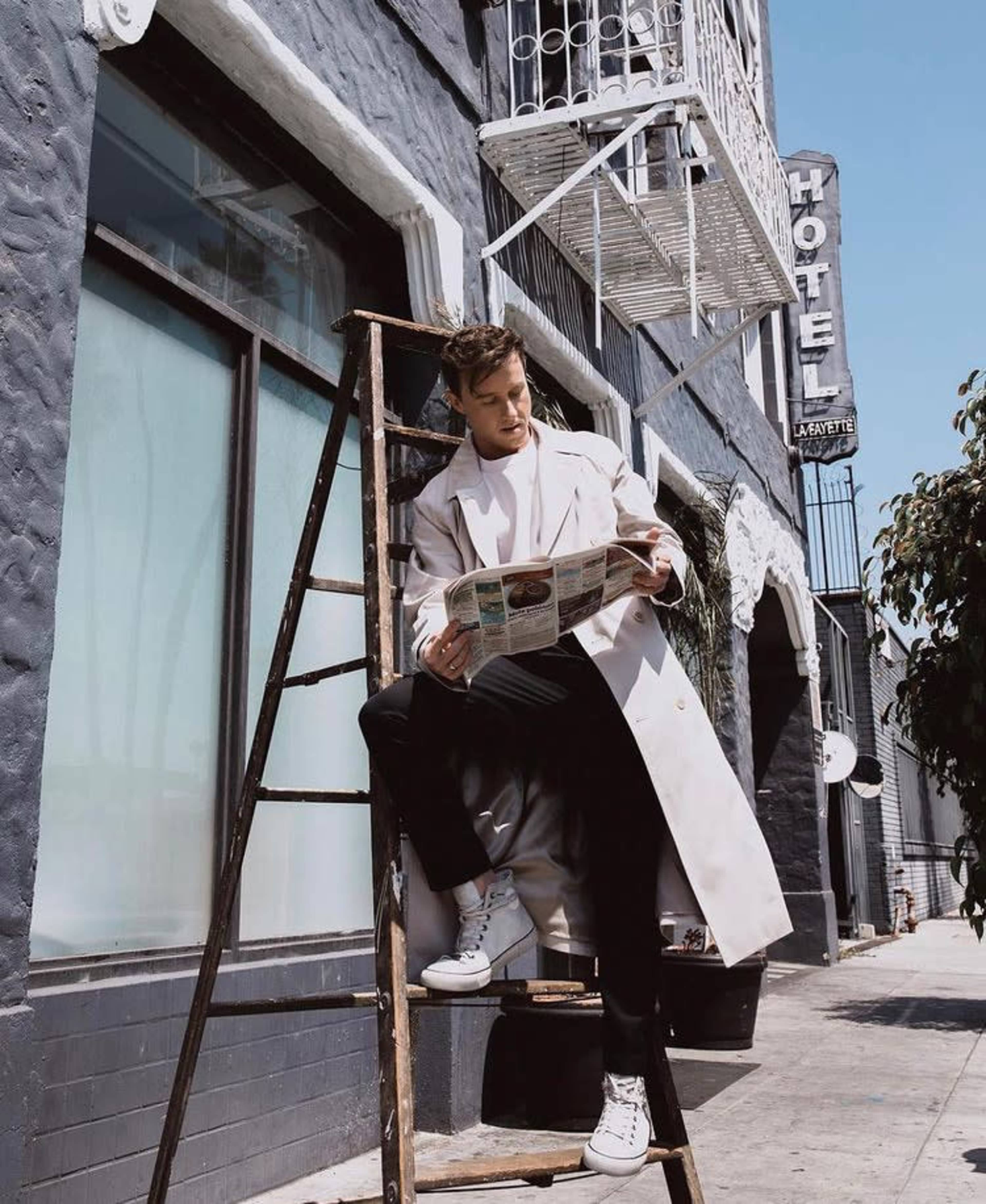 A person sits on a ladder reading a newspaper in front of a building with a "Hotel" sign.