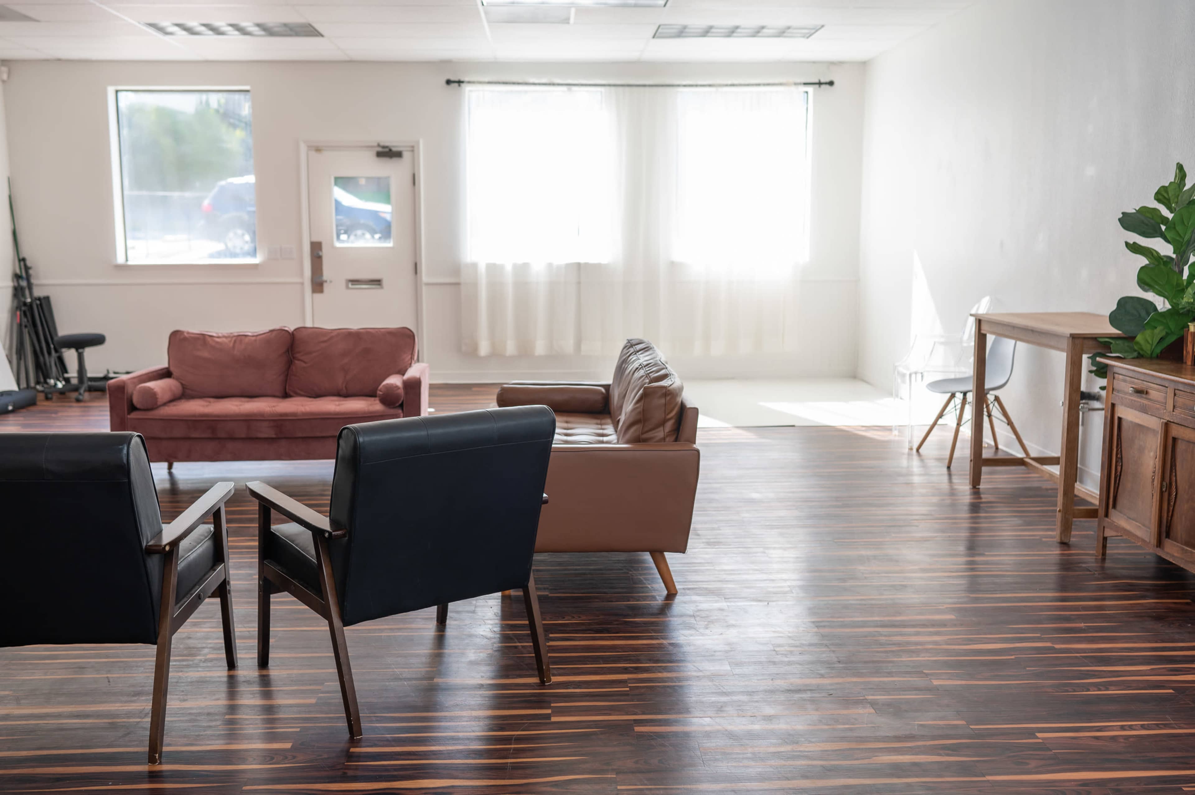 The image shows a spacious room with two sofas, a table, and a window letting in natural light, along with wooden flooring and minimal furnishings.