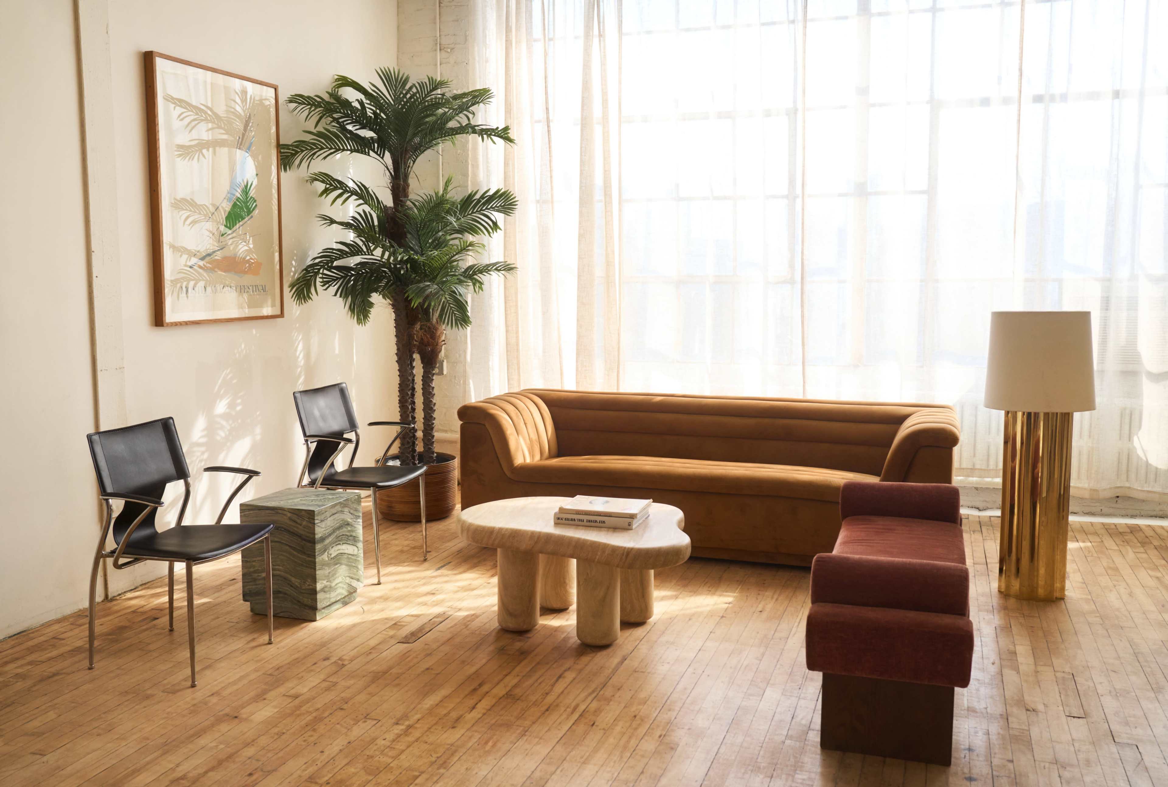 The image shows a bright, modern living room with a brown velvet sofa, two black chairs, a round coffee table, a green stone side table, and a tall lamp, all illuminated by natural light from large windows.