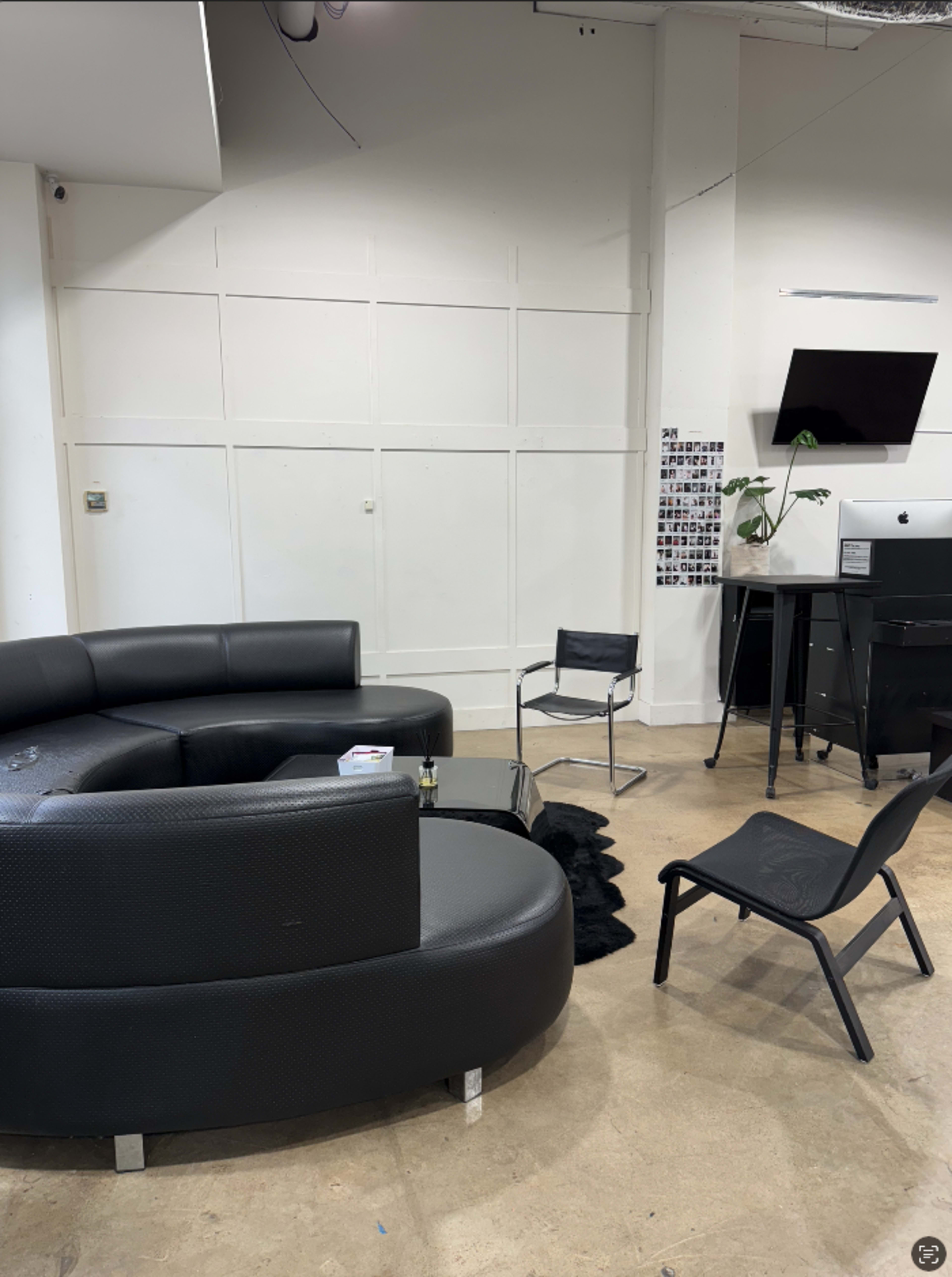 The image shows a modern lounge area with a black circular sofa, a small black chair, a desk with a computer, and minimalist decor against a white wall.