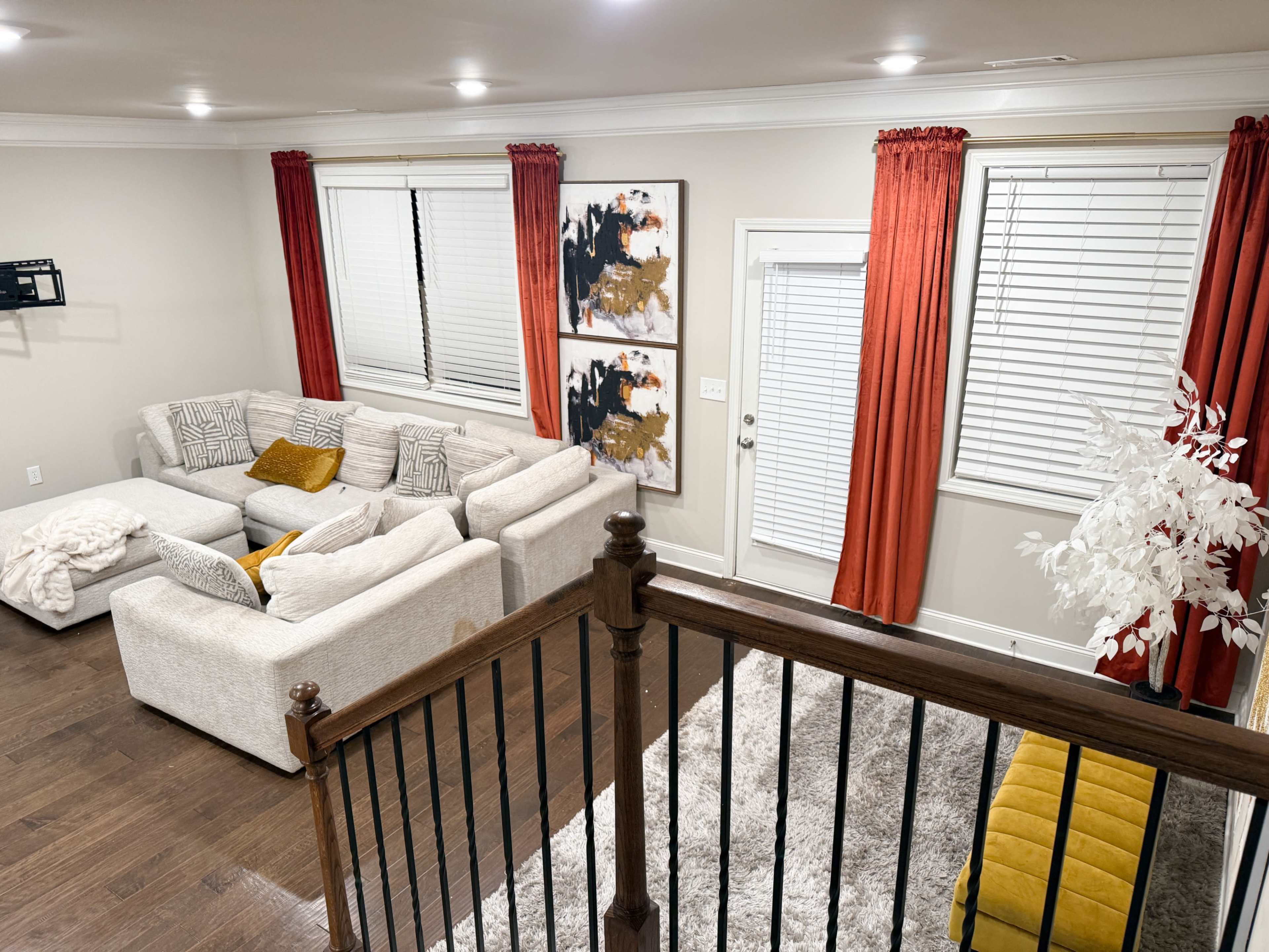 A spacious living room features a large sectional sofa, decorative artwork on the walls, bright curtains over the windows, and a staircase in the foreground.
