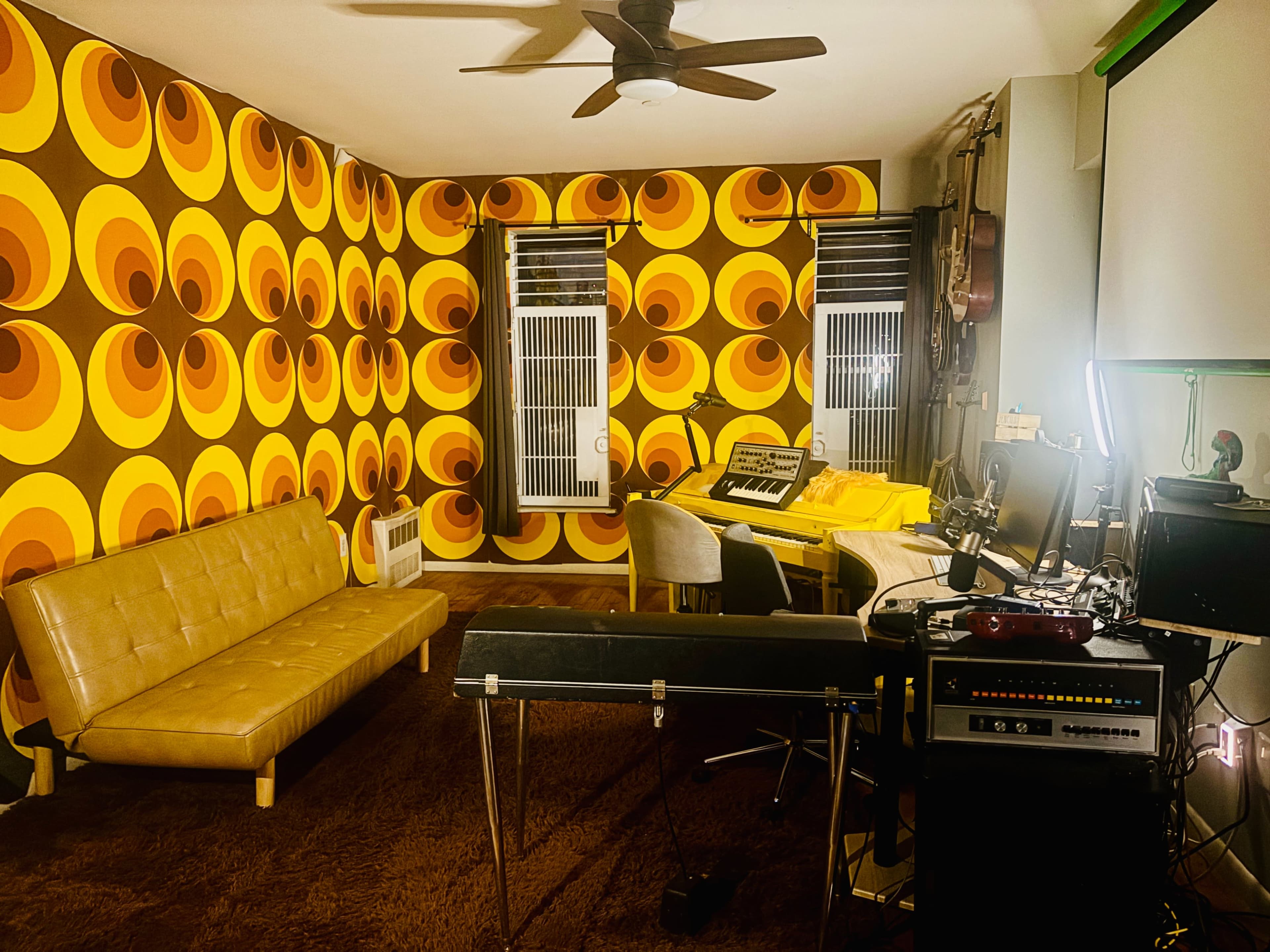 The room features retro circular-patterned wallpaper, a yellow sofa, a keyboard on a table, and various music equipment.
