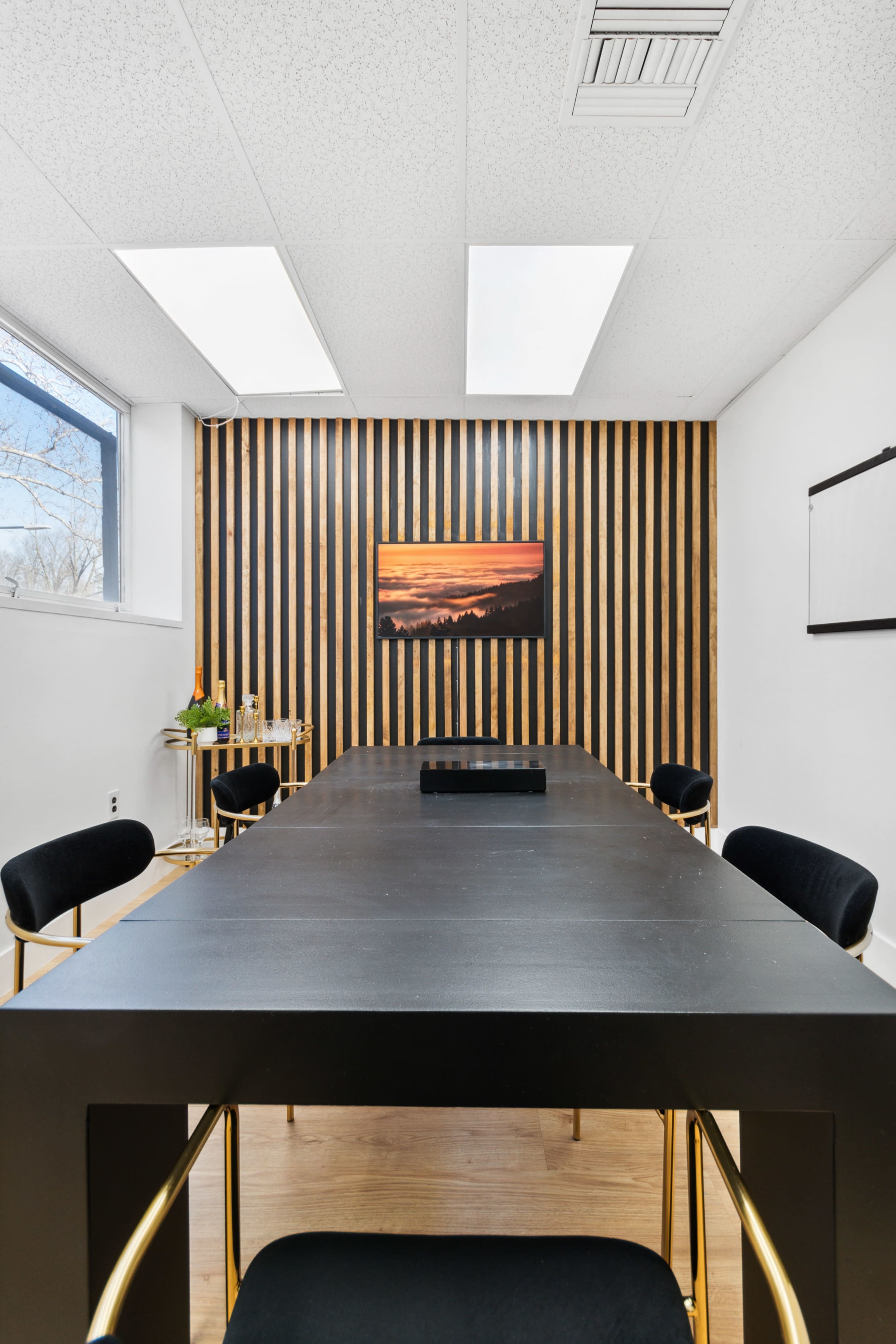 A modern conference room features a long black table surrounded by gold-accented chairs, with a wooden striped accent wall and a landscape painting.