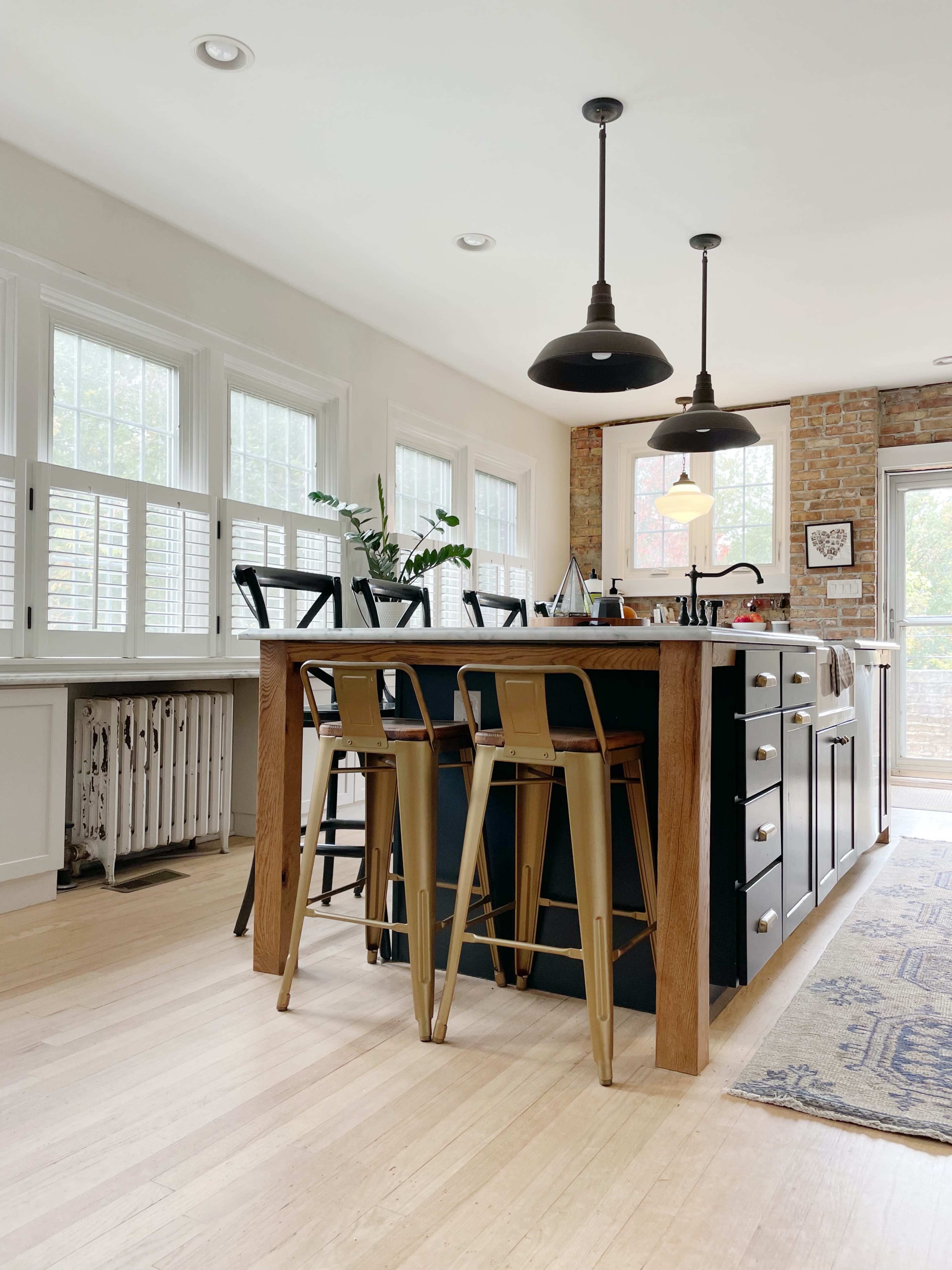 A modern kitchen features a wooden island with metal stools, large windows allowing natural light, and an exposed brick wall.
