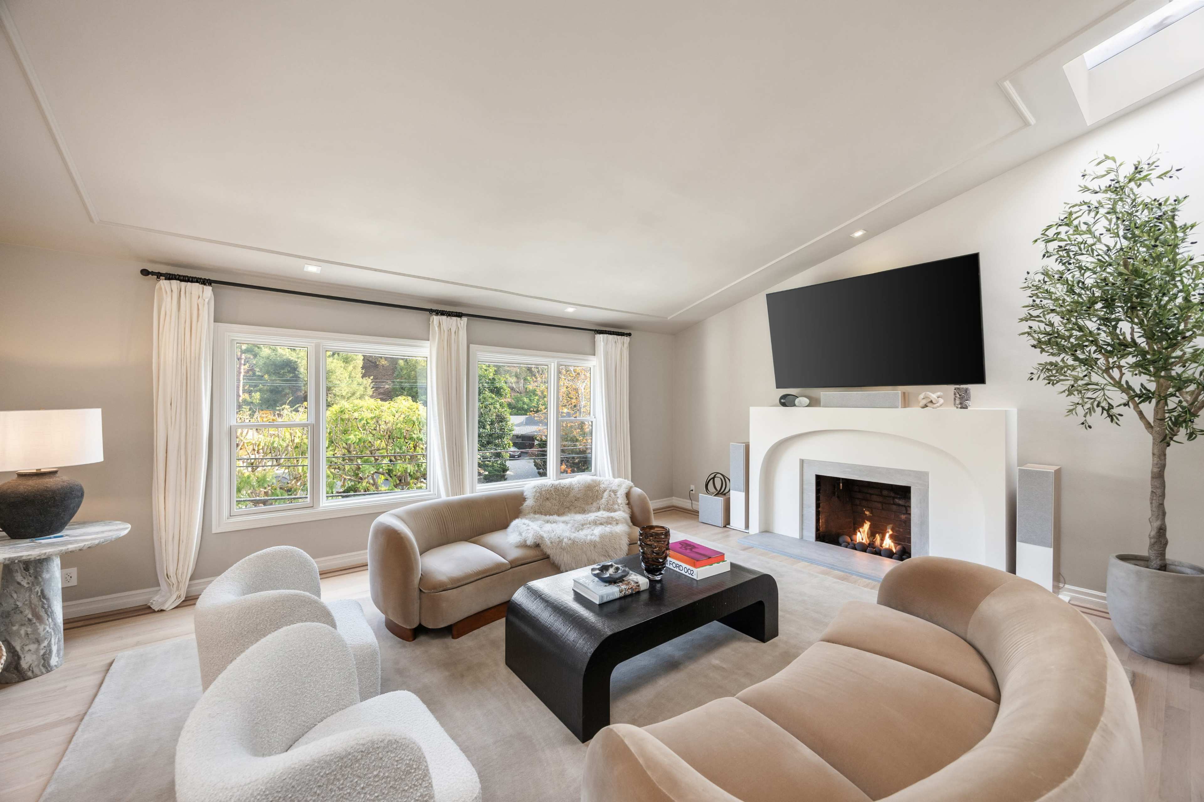 The image shows a modern living room featuring two curved sofas, a black coffee table, a flat-screen TV mounted above a white fireplace, and large windows overlooking greenery.