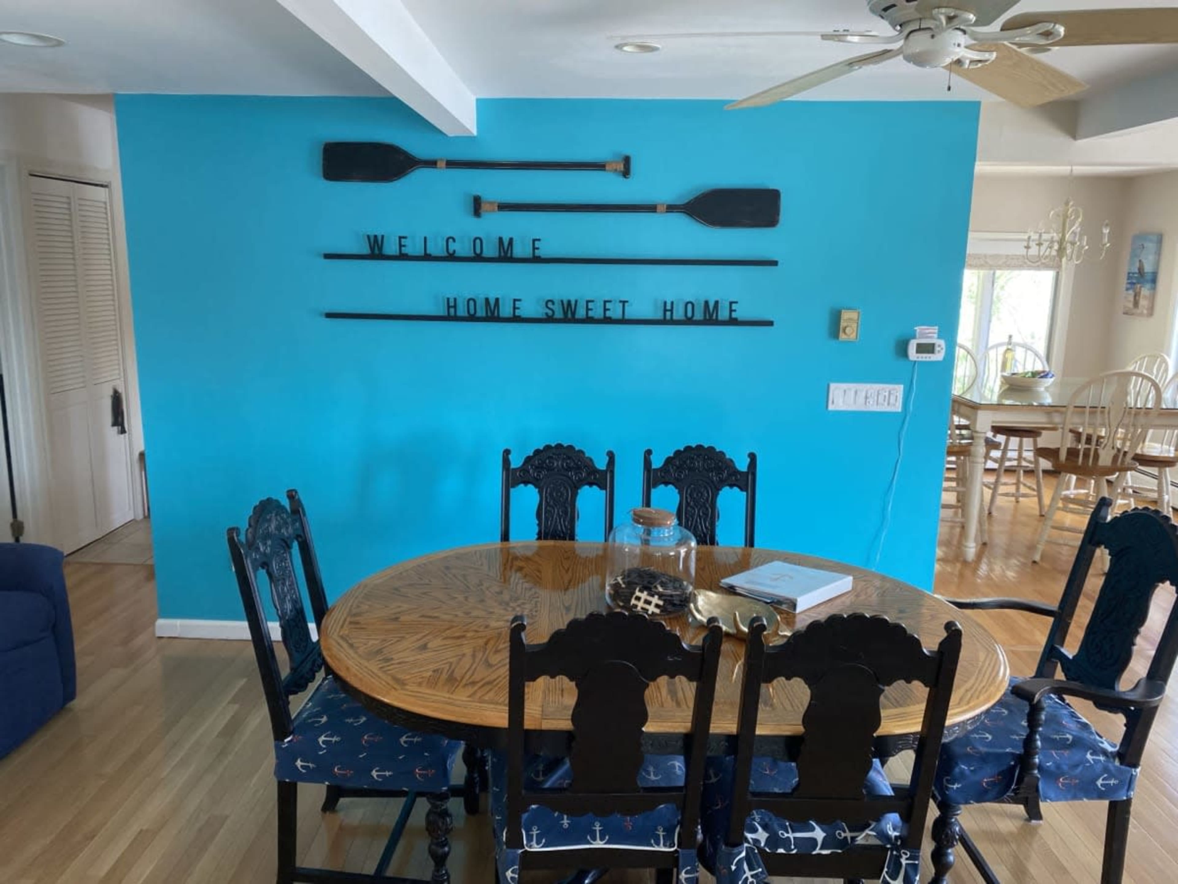 A wooden dining table with eight dark chairs, decorated with a vase and book, sits against a bright blue wall featuring paddles and a welcome sign.