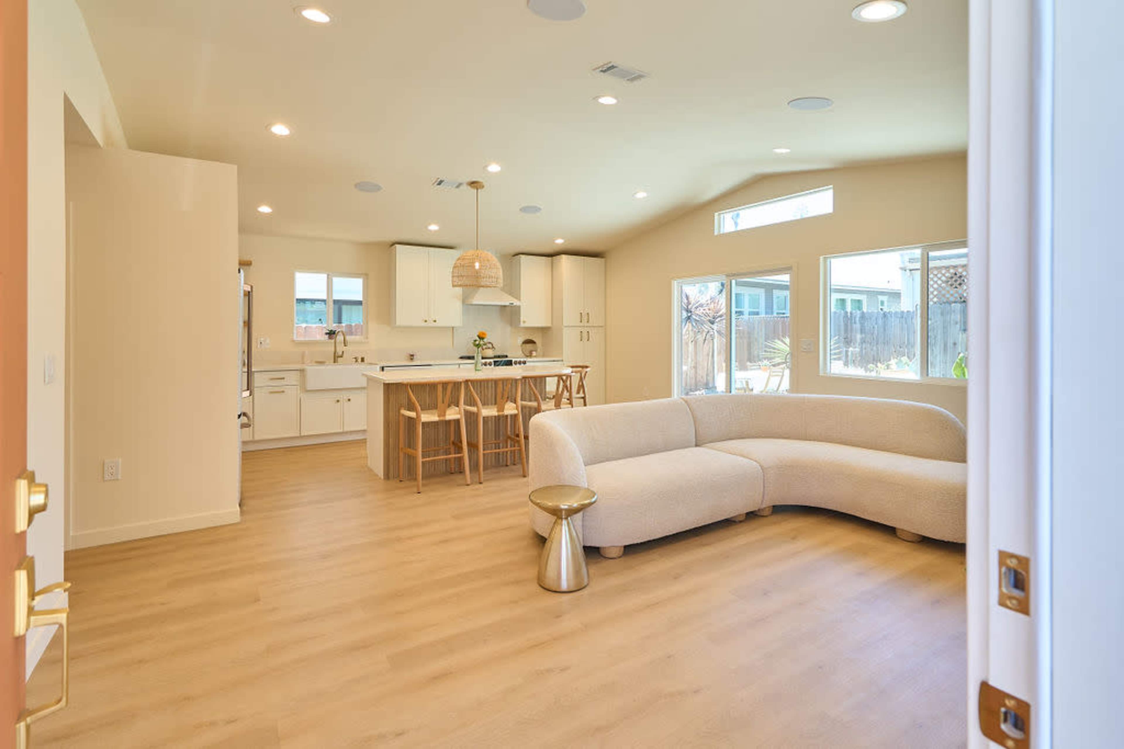 The image shows a bright, modern open-concept living area that features a curved sofa, a kitchen with an island, and large windows allowing natural light to fill the space.
