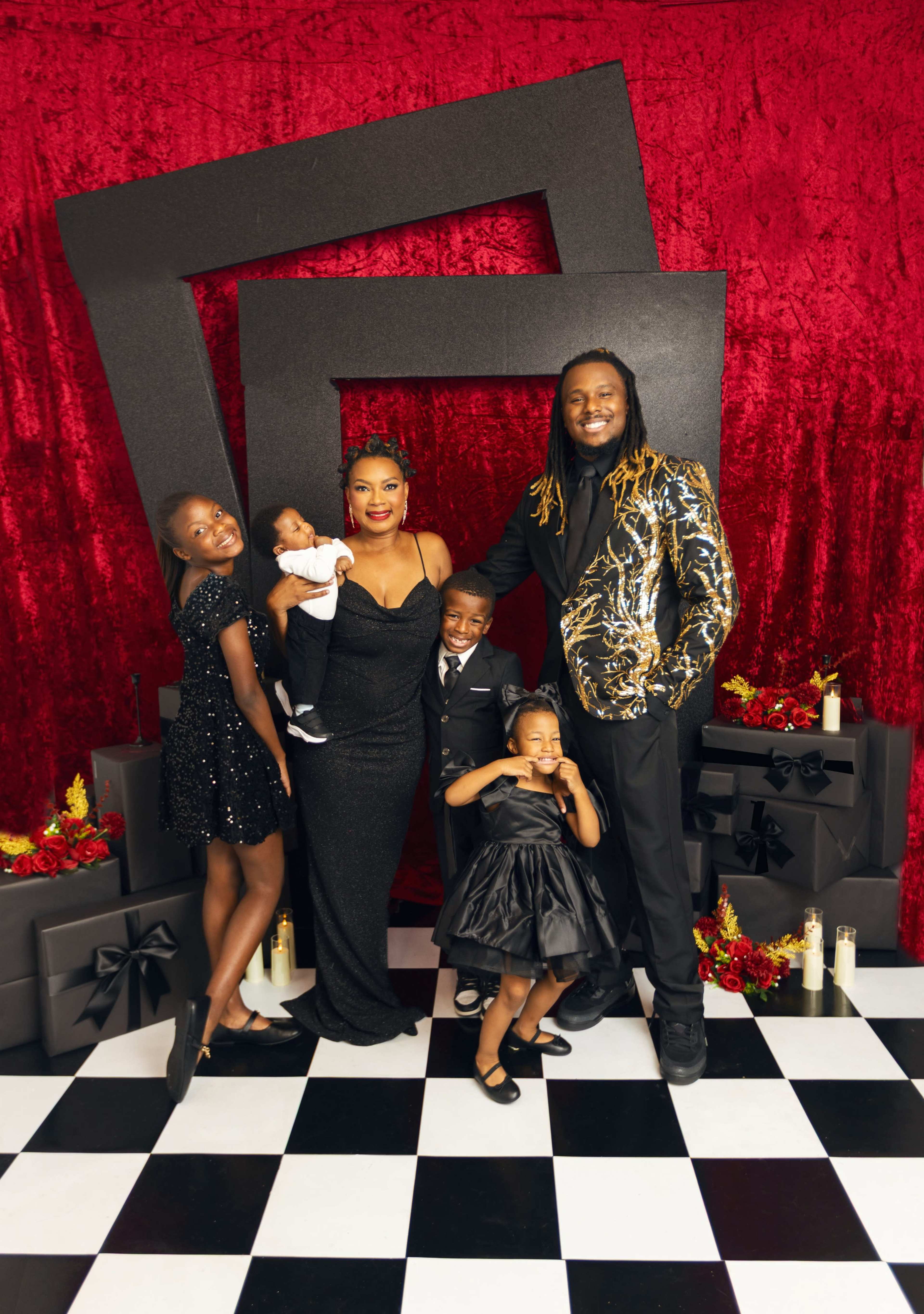 A family poses for a photo in front of a red and black backdrop with geometric shapes, surrounded by decorative flowers and candles on a black-and-white checkered floor.
