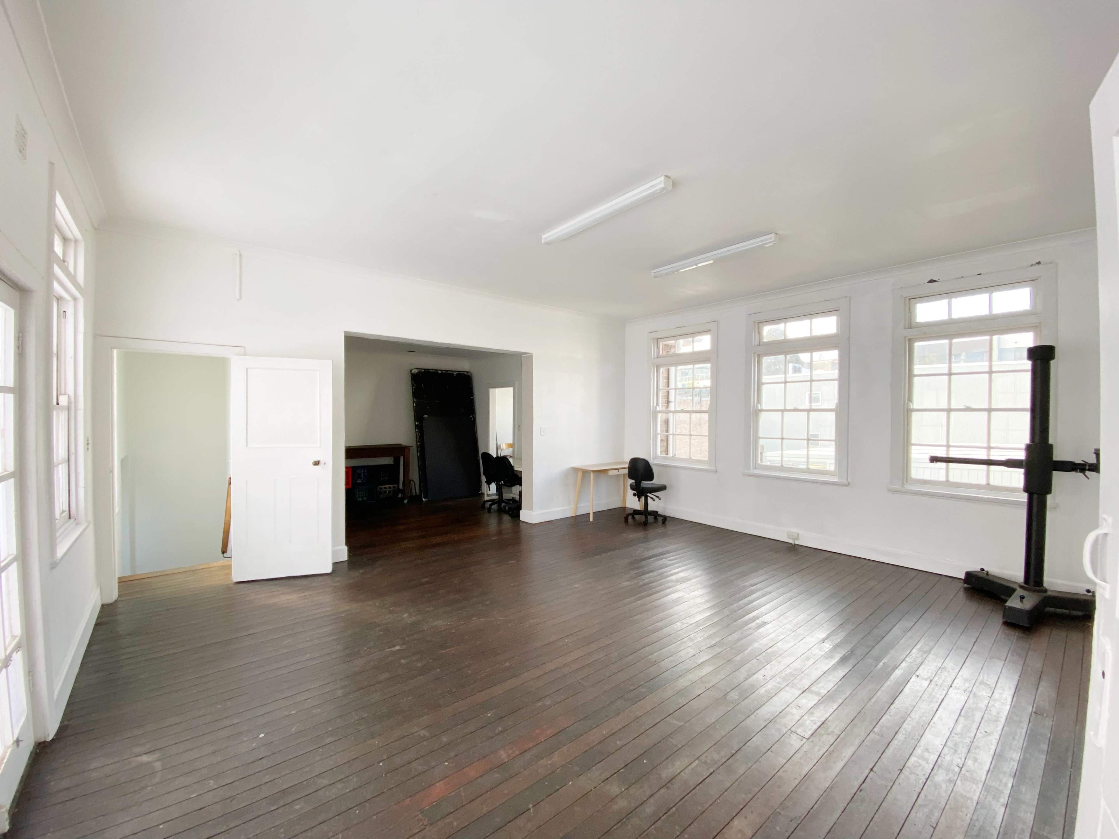A bright, empty room features wooden floors, large windows, and an adjoining space with a desk and chair.