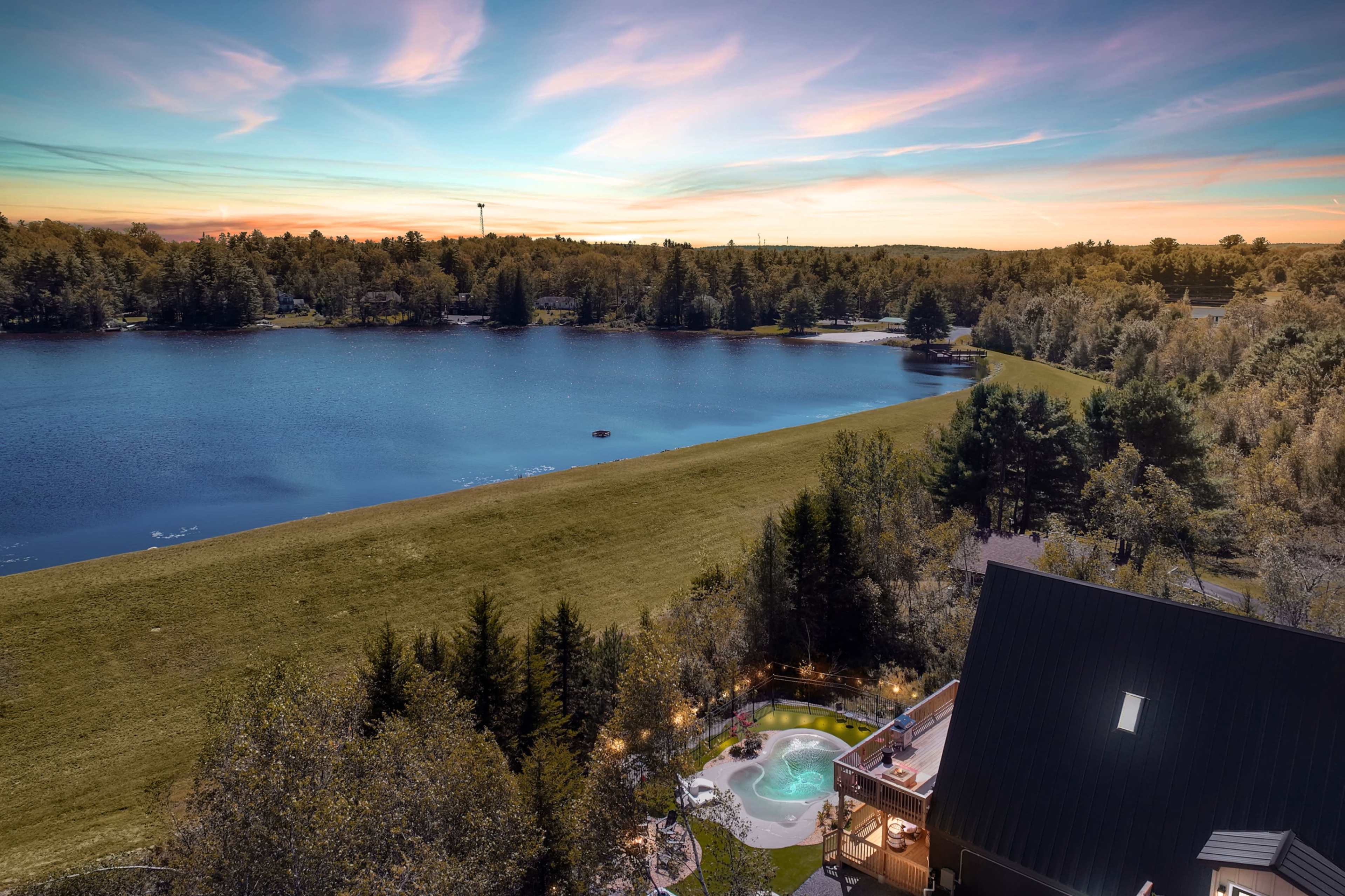 The image shows a serene lakeside scene with a house on the right, a hot tub on a patio, and lush greenery surrounding the calm water under a twilight sky.